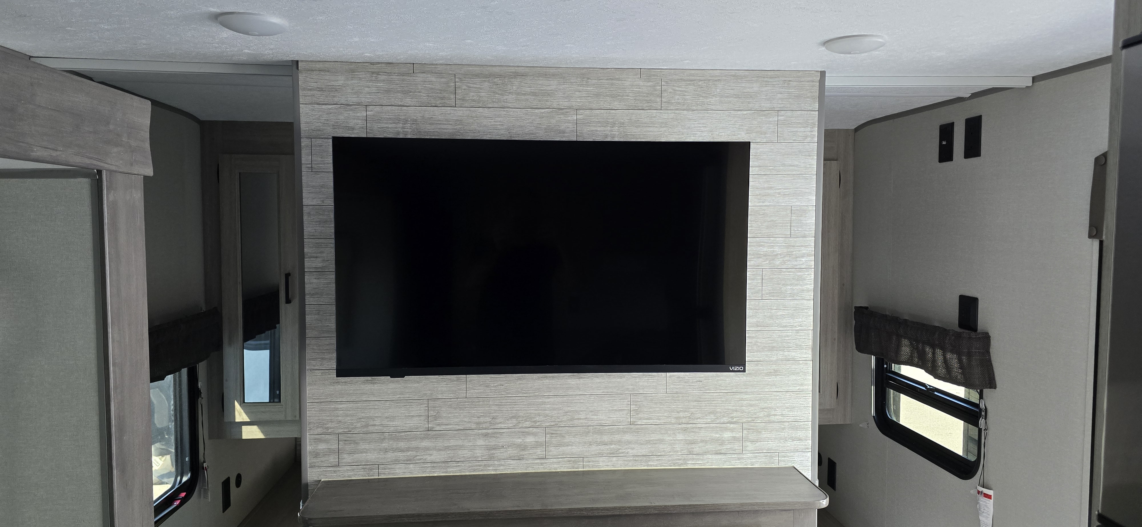 Modern rv rental interior with a large flat-screen TV on a gray plank accent wall, showing windows, cabinetry, and light ceiling.. Coachmen Catalina 2025