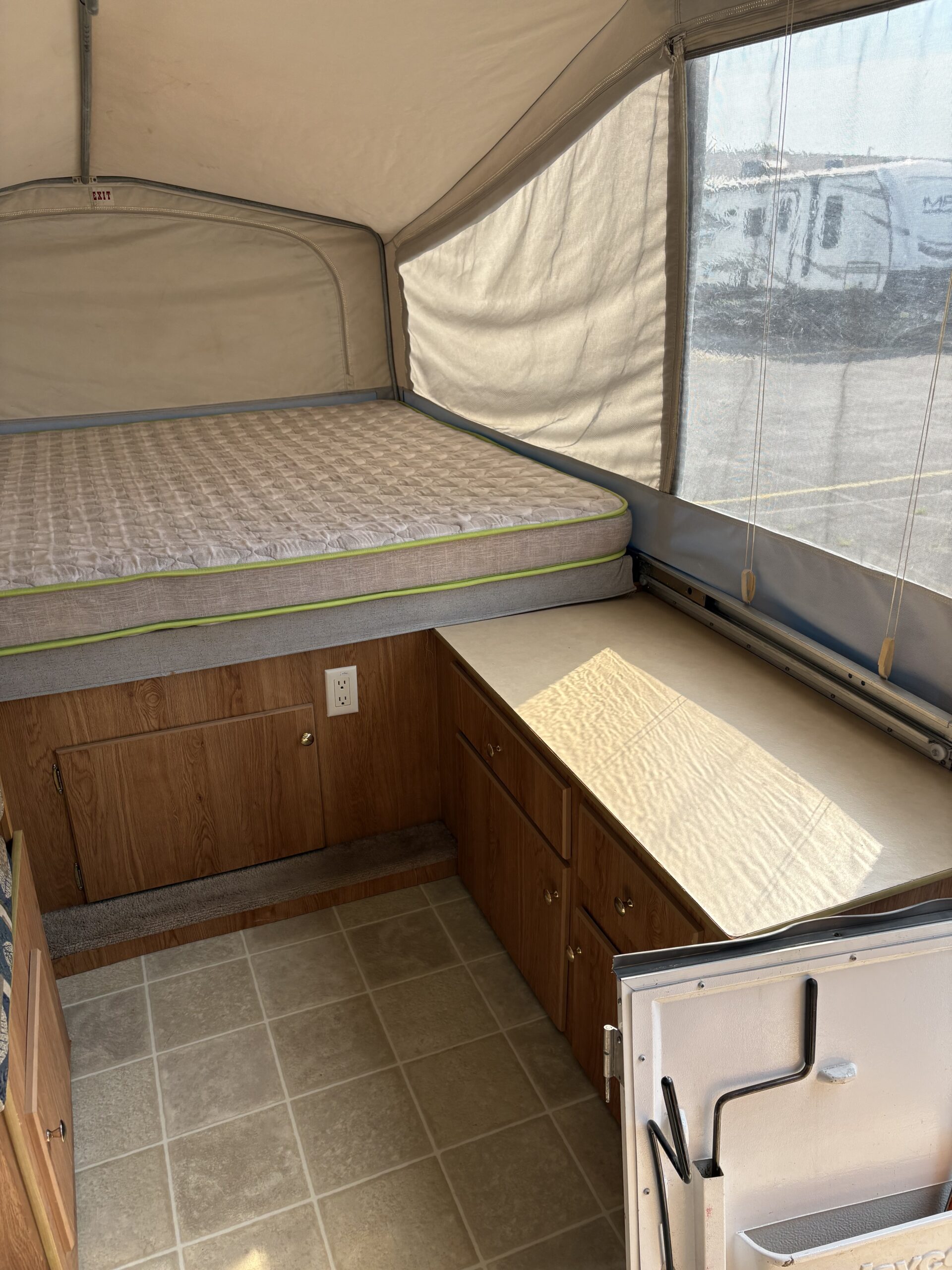 RV rental pop-up camper's spacious interior: comfortable bed, wood-paneled storage, and tiled floor. Natural light from window.. Jayco Jay Series 2006