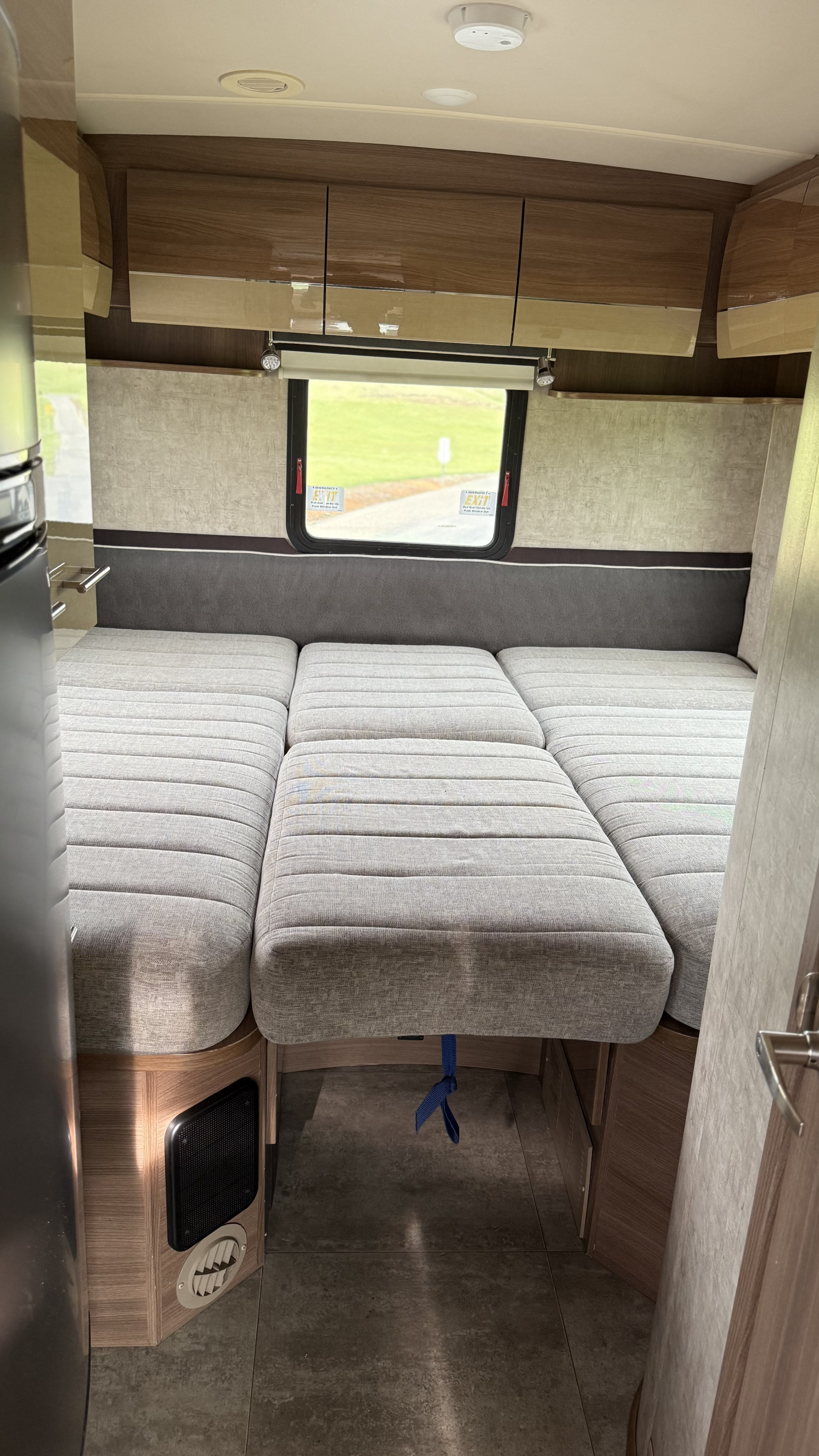 View of an RV rental's spacious interior: light grey bed cushions, wooden overhead cabinets, window, and fridge.. Itasca Navion 2016