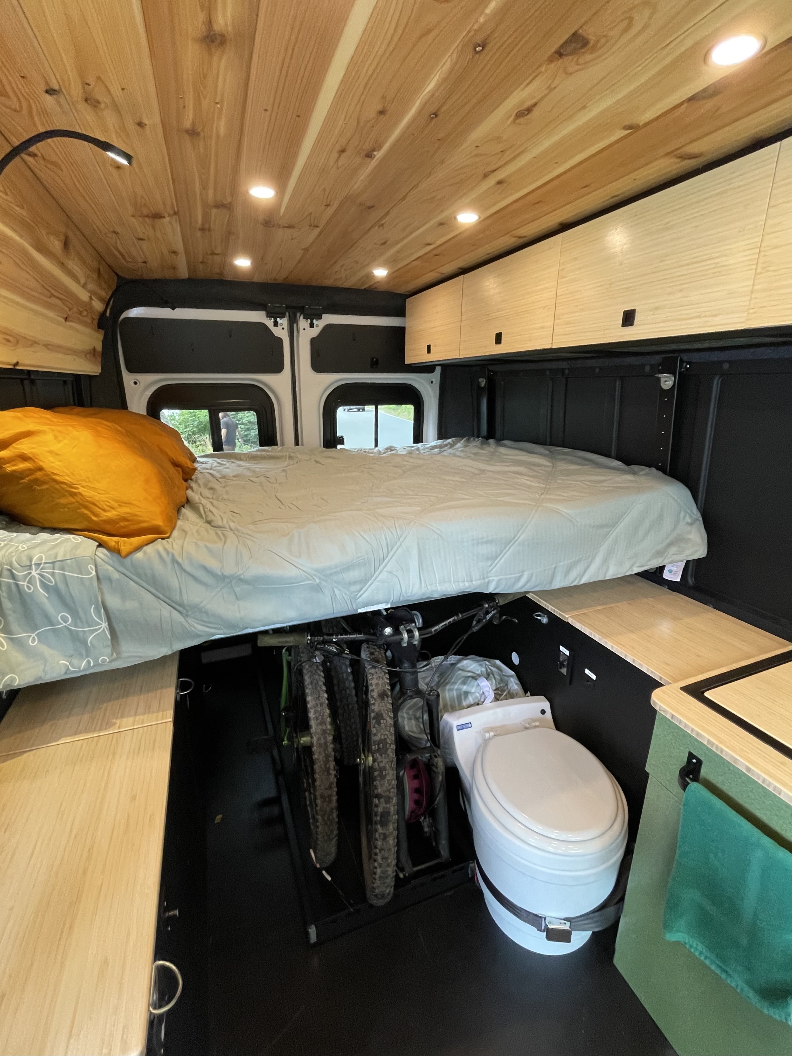 Cozy RV rental interior features a raised bed, wood ceiling, cabinets, mountain bikes, and a portable toilet.. Ram Promaster 2023
