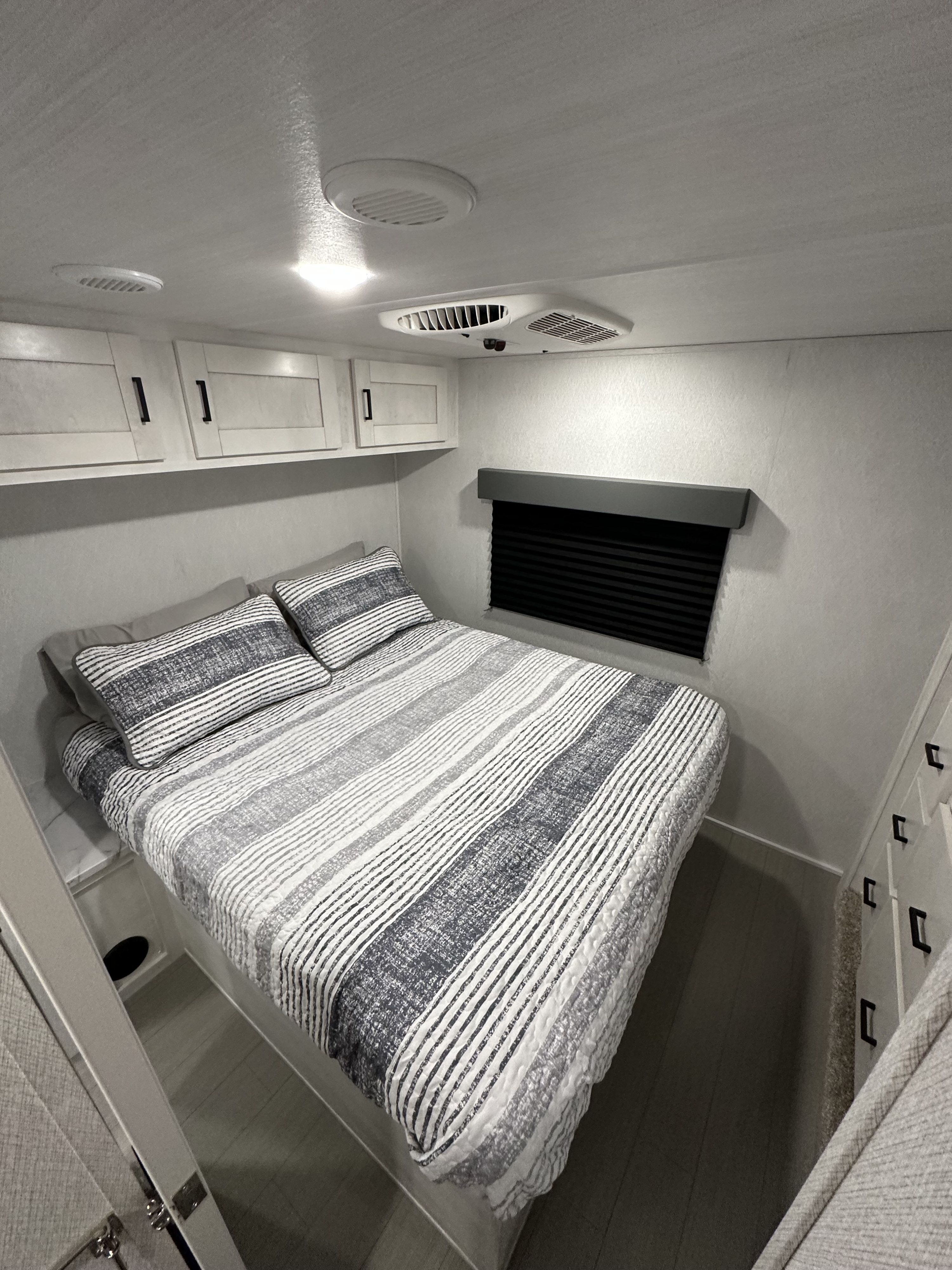 Cozy RV rental bedroom featuring a bed with striped bedding, overhead cabinets, AC unit, and window blind.. East to West Entrada 3100FB 2025