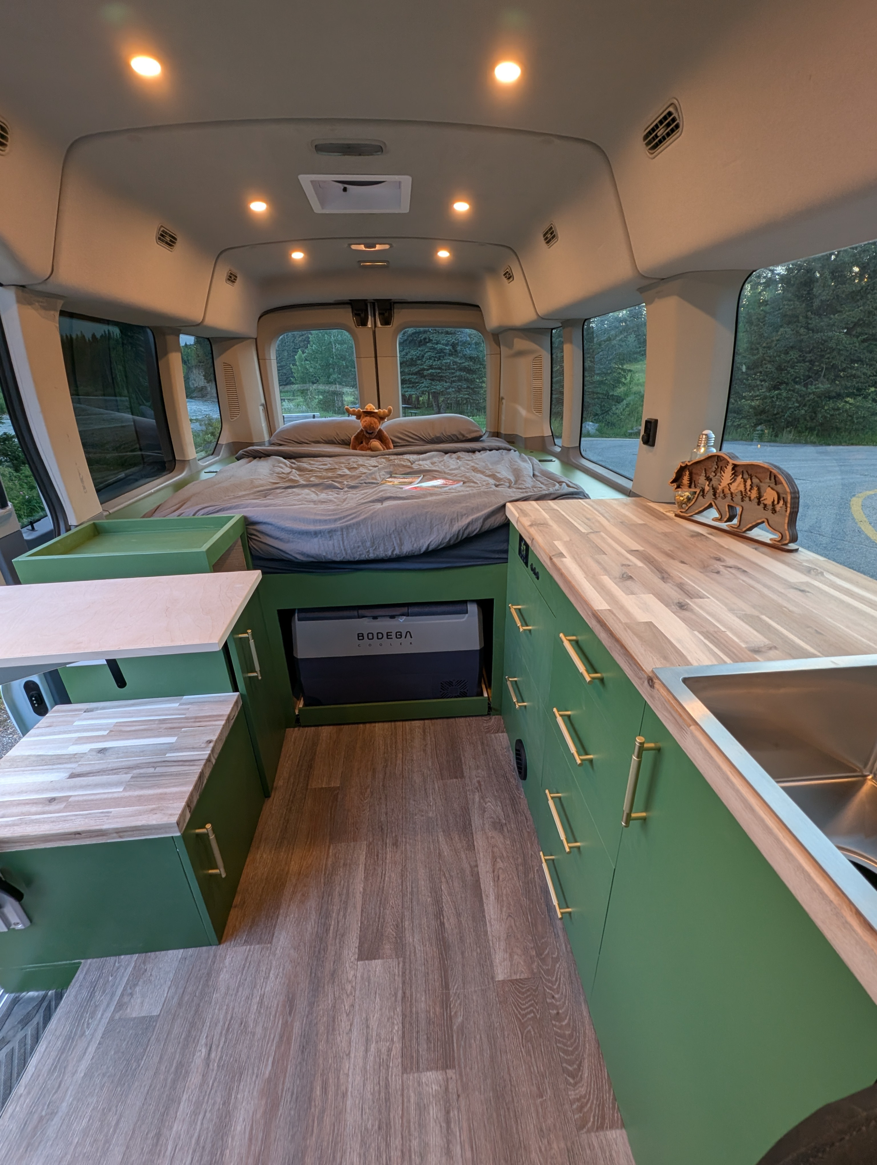 Spacious RV rental interior featuring a cozy bed, green kitchen cabinets with wood counters & sink, and a pull-out cooler. Perfect for your next road trip adventure!. Ford Transit 2017
