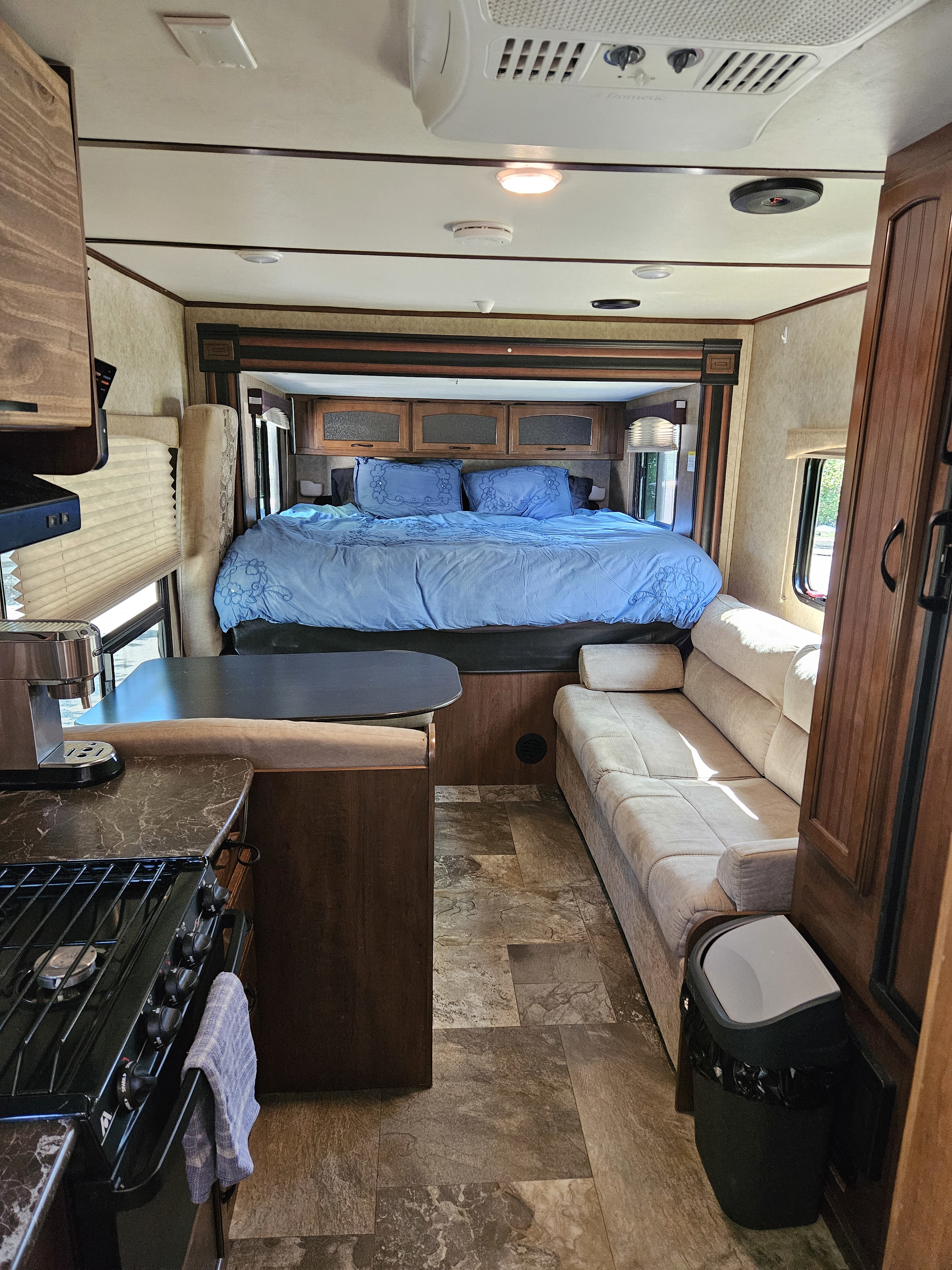 Bright RV rental interior. Features bedroom with blue bedding, comfy living room couch, & kitchen area with stove/coffee maker.. Jayco X213 2016