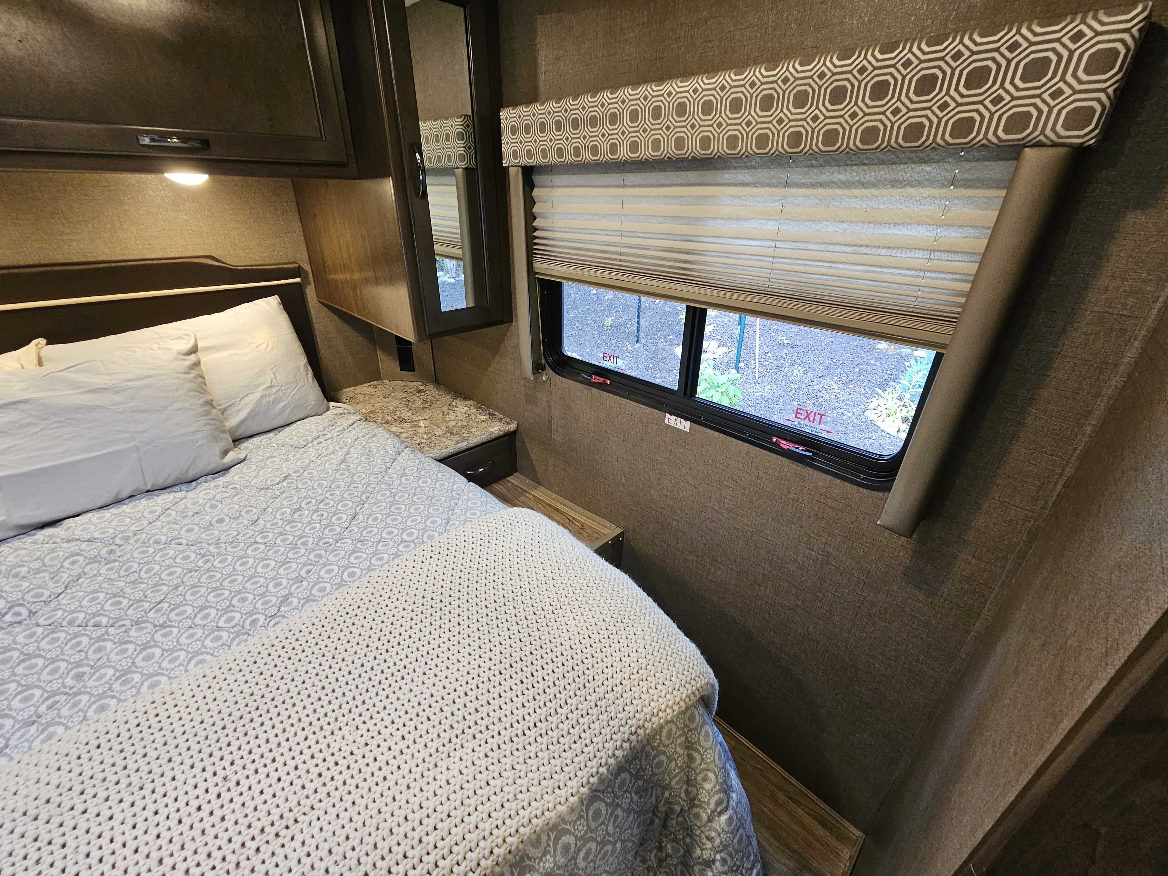This RV rental bedroom includes a cozy bed with white linens, overhead storage, and a window with blinds showing an emergency exit.. Thor Four Winds 2019