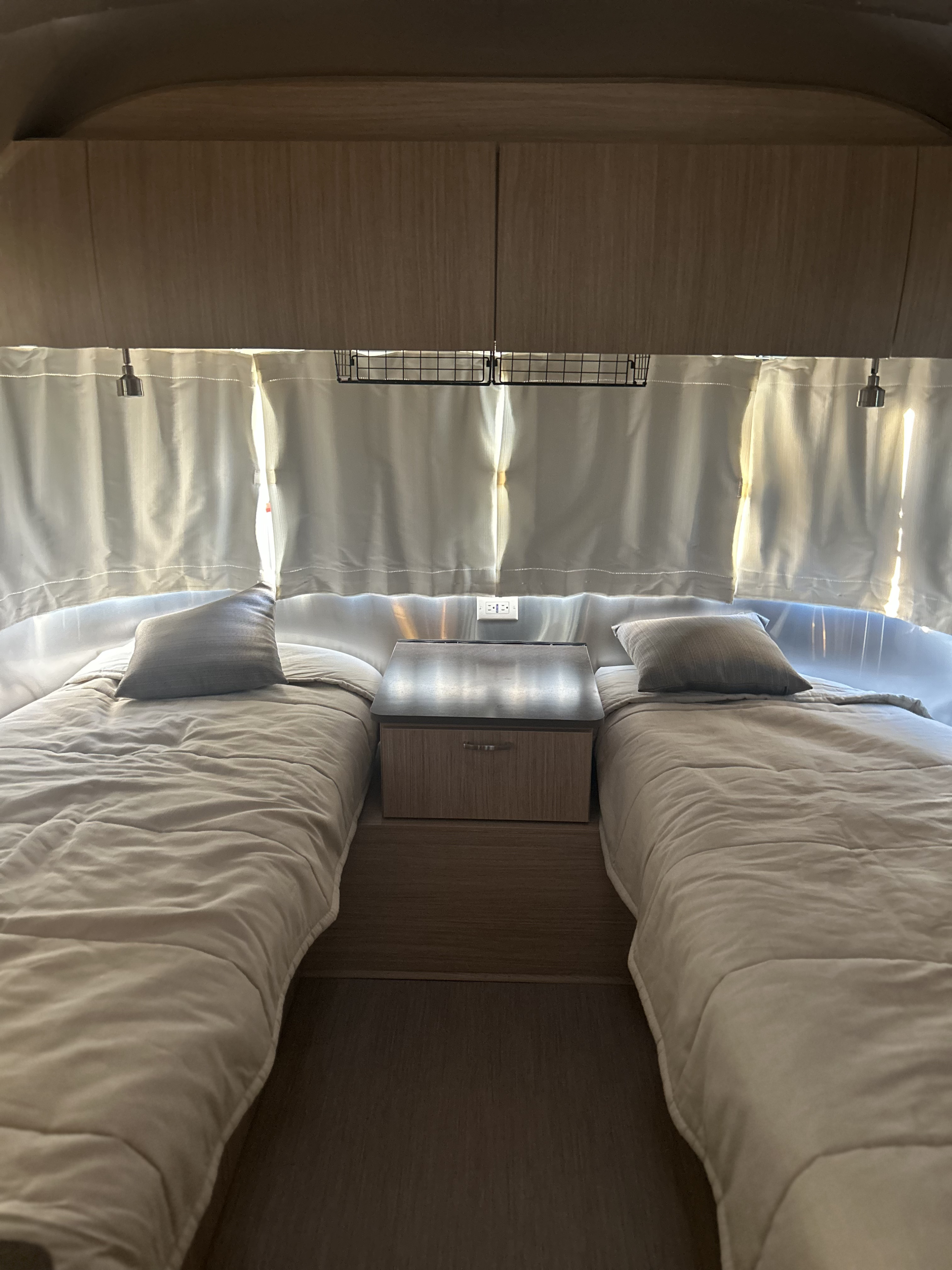 This RV rental bedroom features cozy twin beds, a central nightstand, and bright window curtains. Ideal for mobile comfort.. Airstream 26RB TWIN 2018