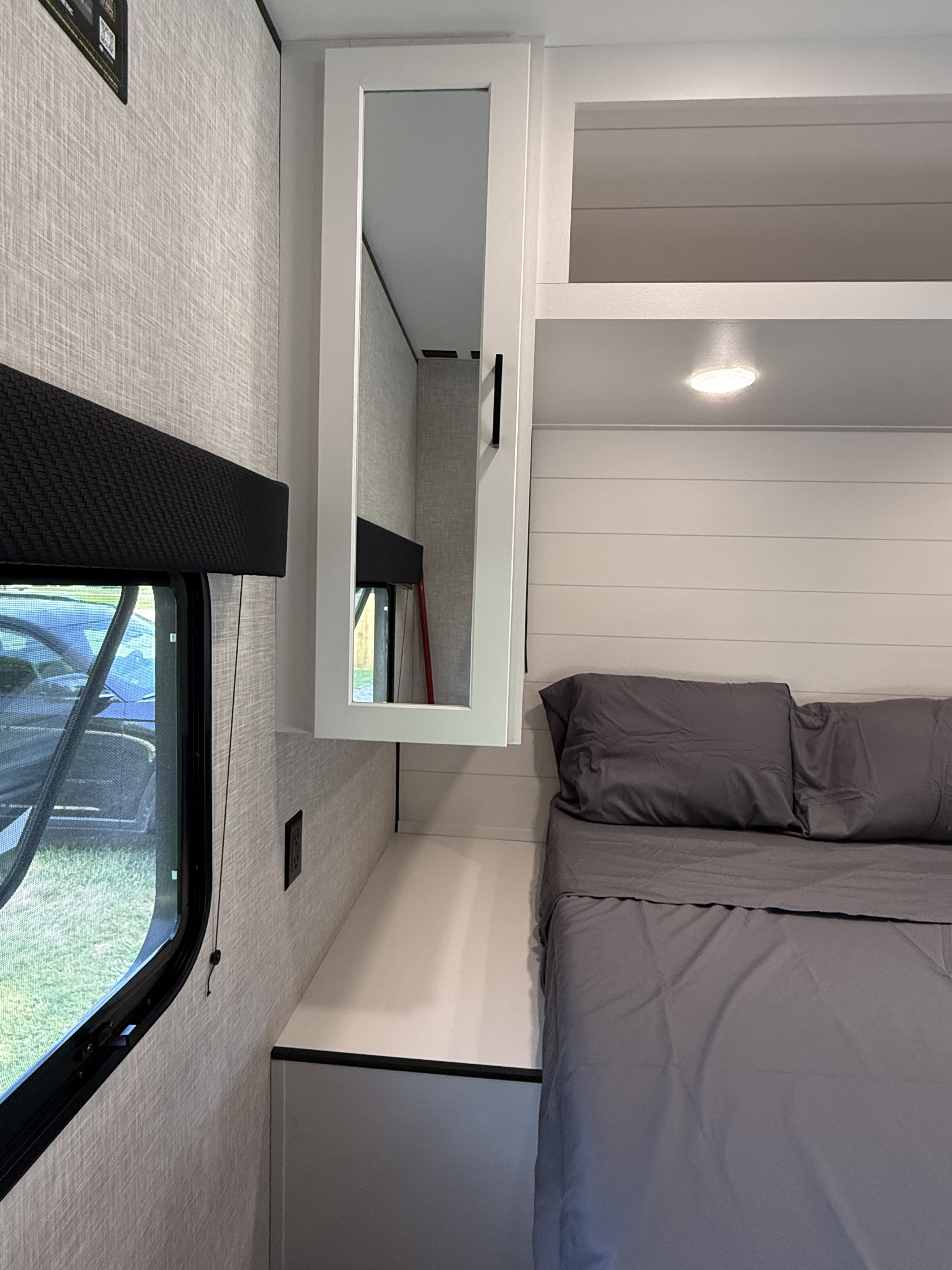 Compact RV rental bedroom showing a gray bed, mirrored cabinet, window, and wall outlet. Cozy and functional space.. Jayco Jay Flight 2025