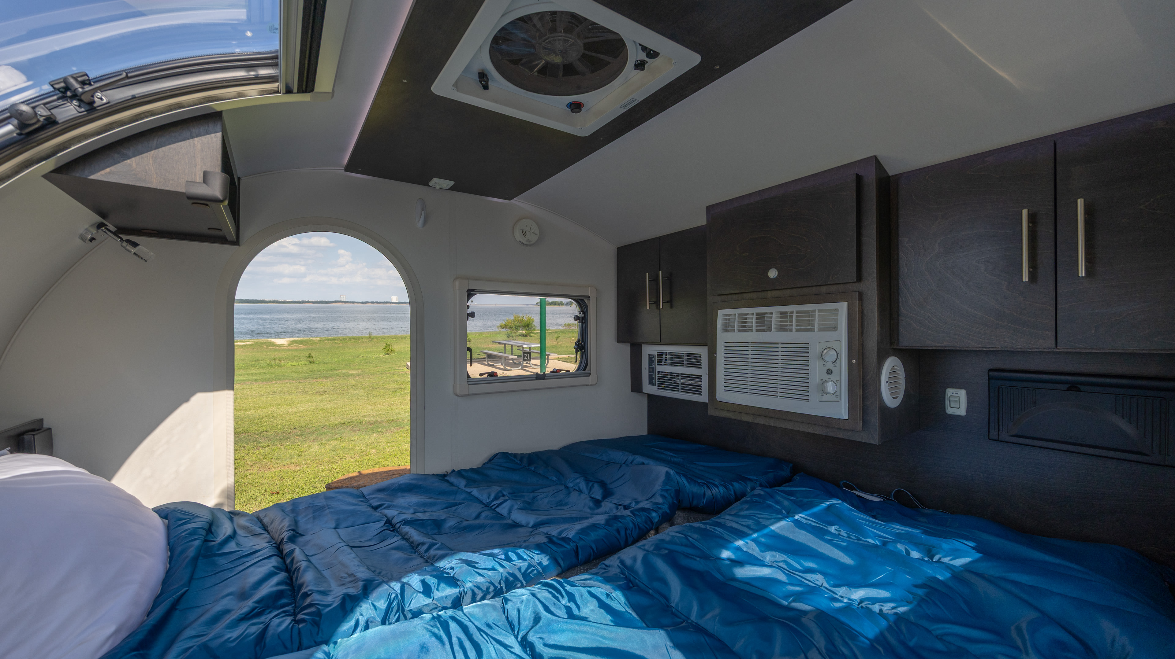 Inside a compact RV rental: two beds, AC units, and dark wood cabinets. Offers a scenic lake view from the entrance.. nuCamp T@G 2022