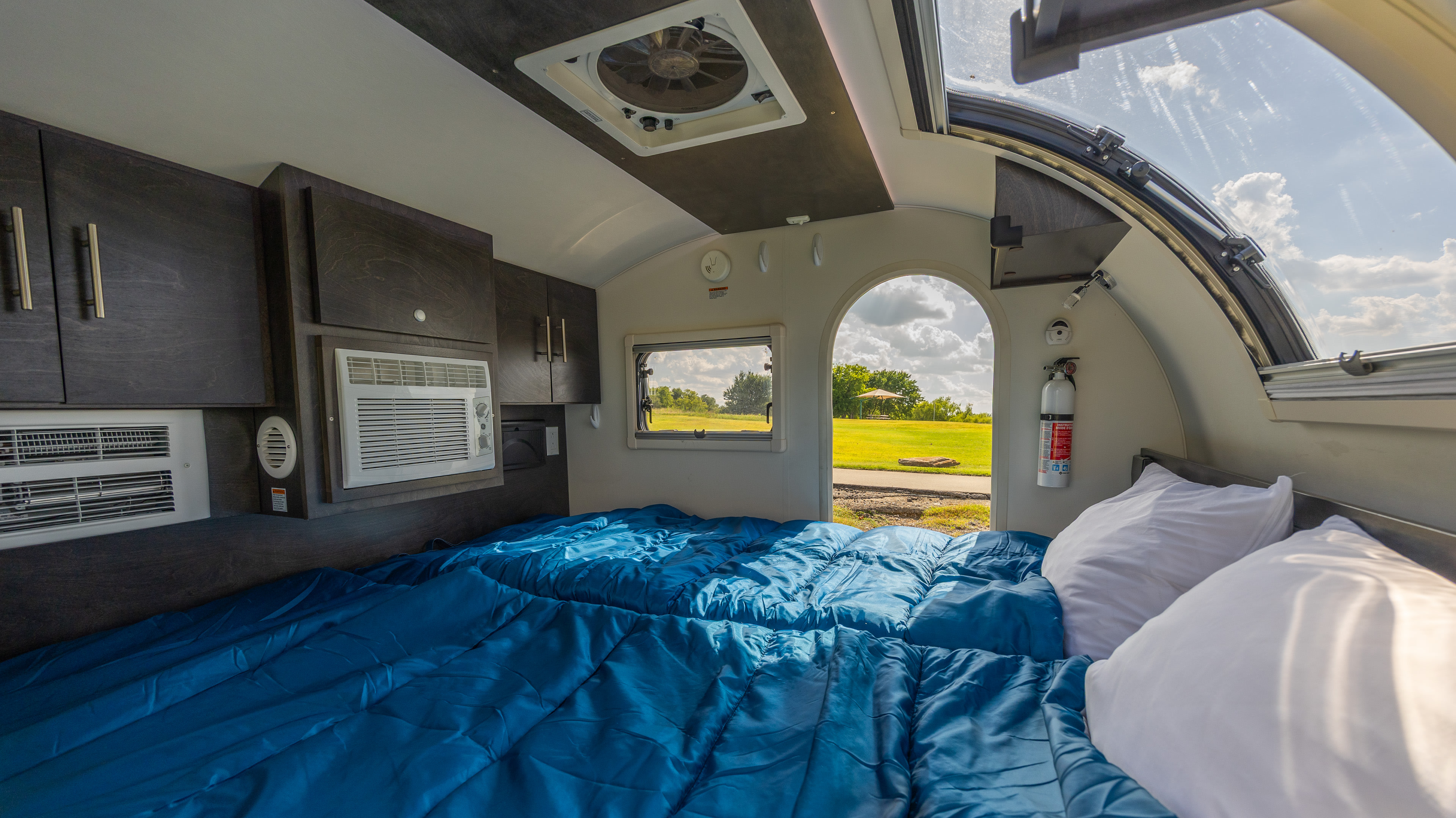Inside a cozy RV rental: two beds with blue covers, AC units, and a large window view of nature.. nuCamp T@G 2022