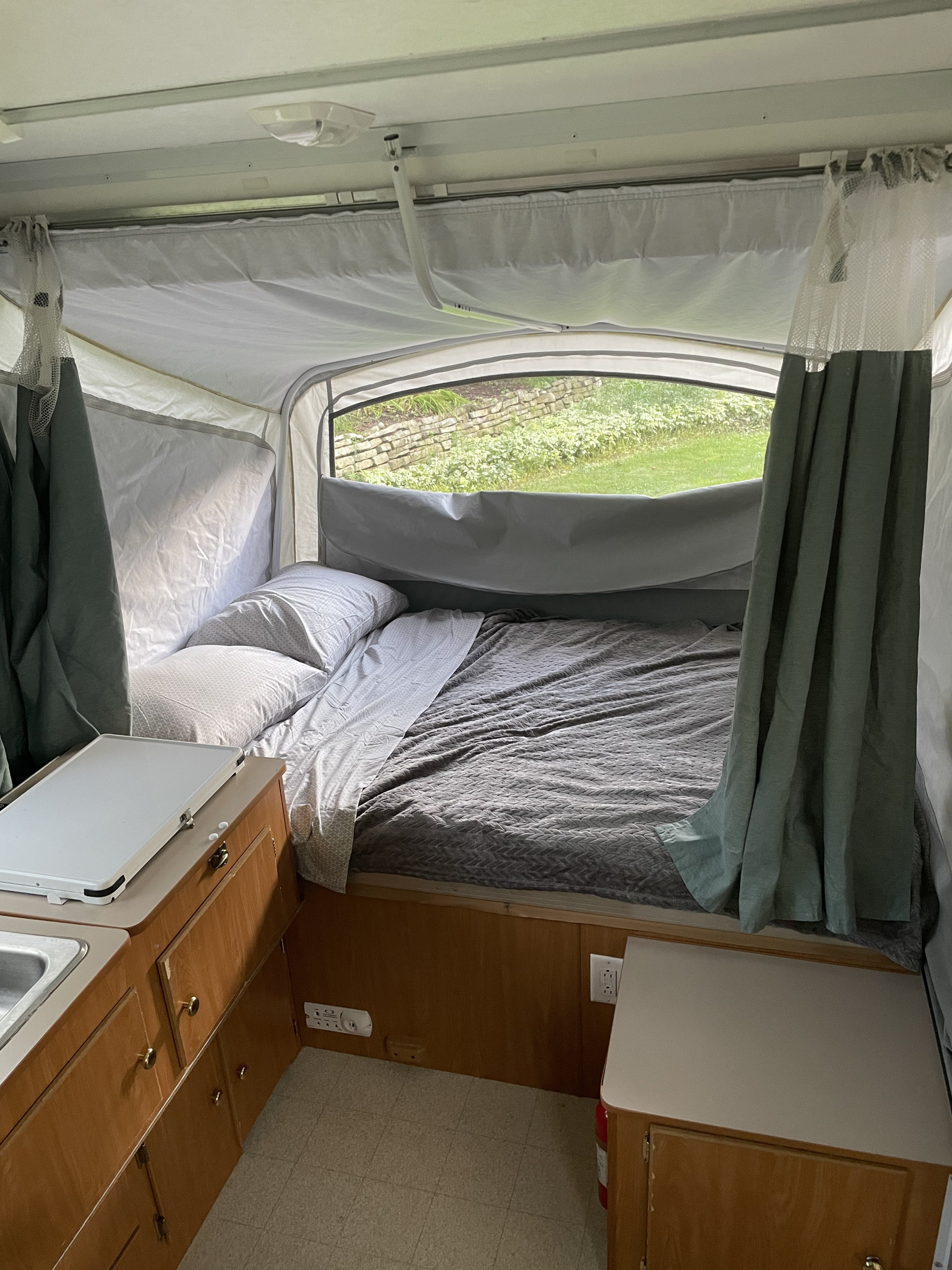 Interior of an RV rental pop-up camper featuring a bed with bedding, compact kitchen, and a window view of greenery.. Coleman Taos 2002