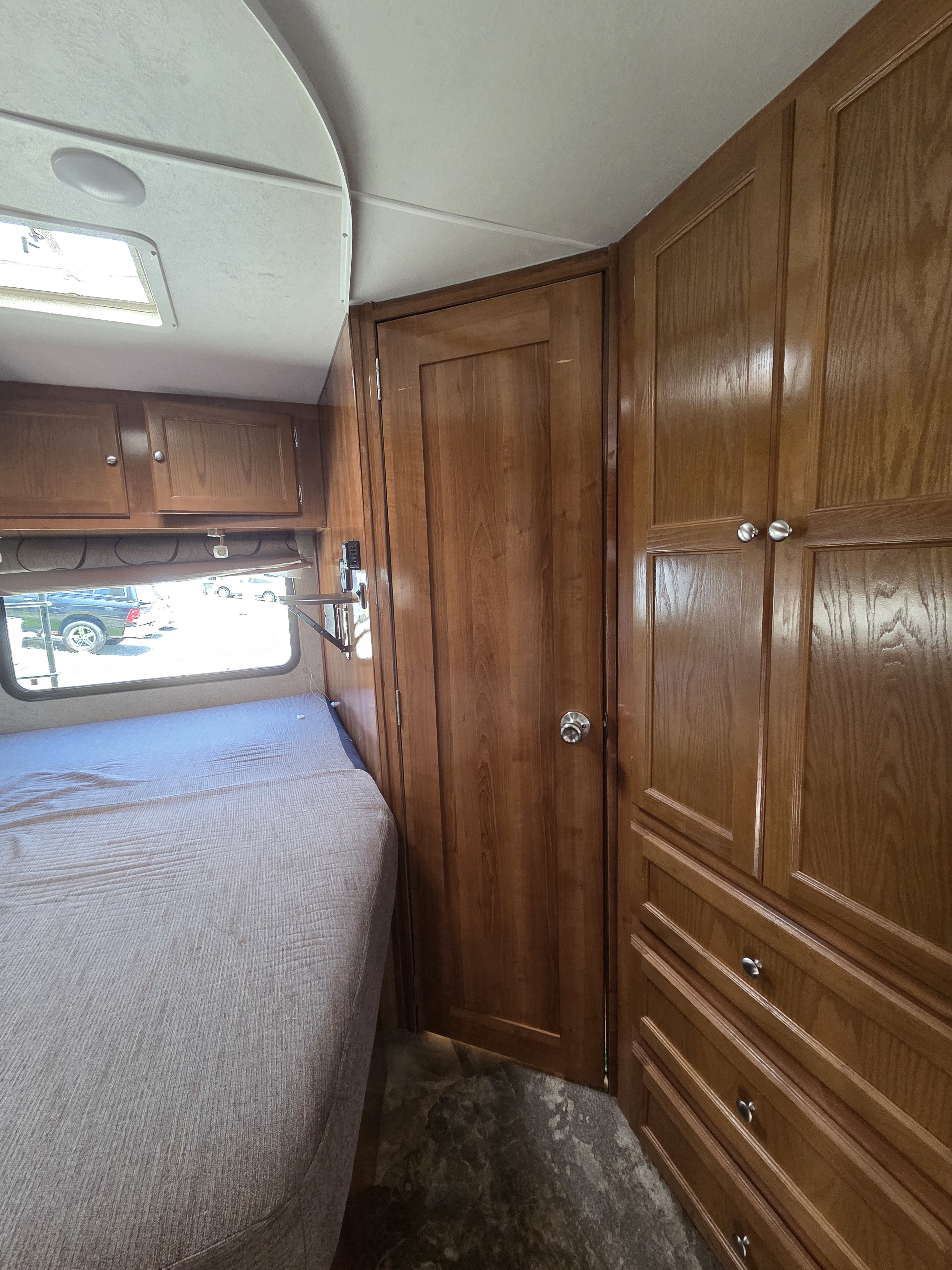 RV rental bedroom showing bed, window, overhead cabinets, a door, and a built-in wooden wardrobe for storage.. Forest River Coachmen Leprechaun 2019