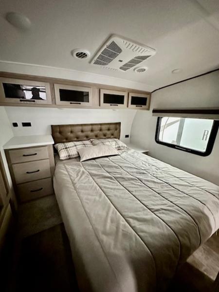 A comfortable RV rental bedroom featuring a large bed, nightstand with drawers, overhead cabinets, and ceiling AC unit.. Forest River Rockwood Signature 2024