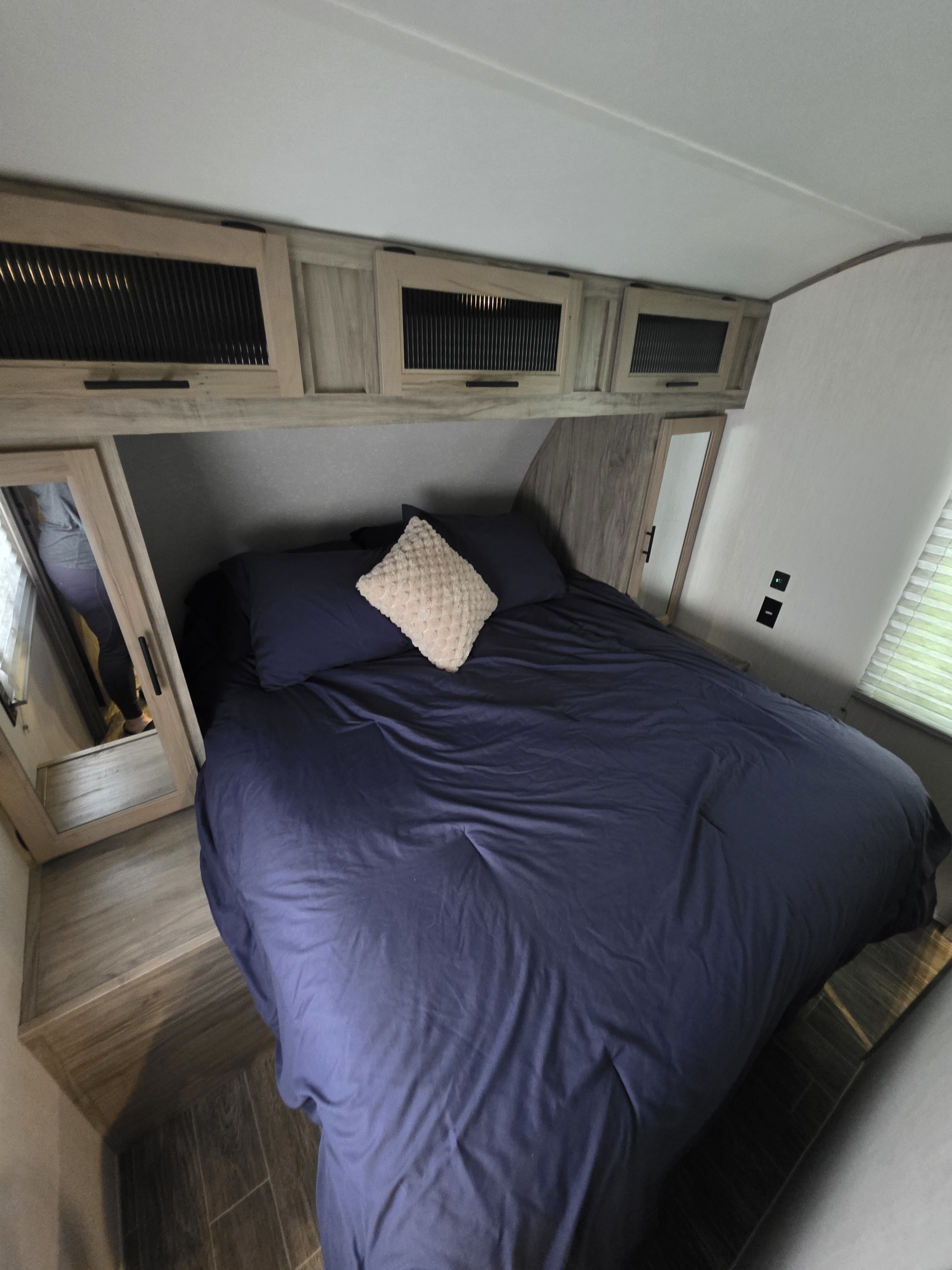 Cozy RV rental bedroom: large bed with navy linens, light pillow, and smart overhead storage. Person reflected in mirror.. Forest River Cherokee Alpha Wolf 2021