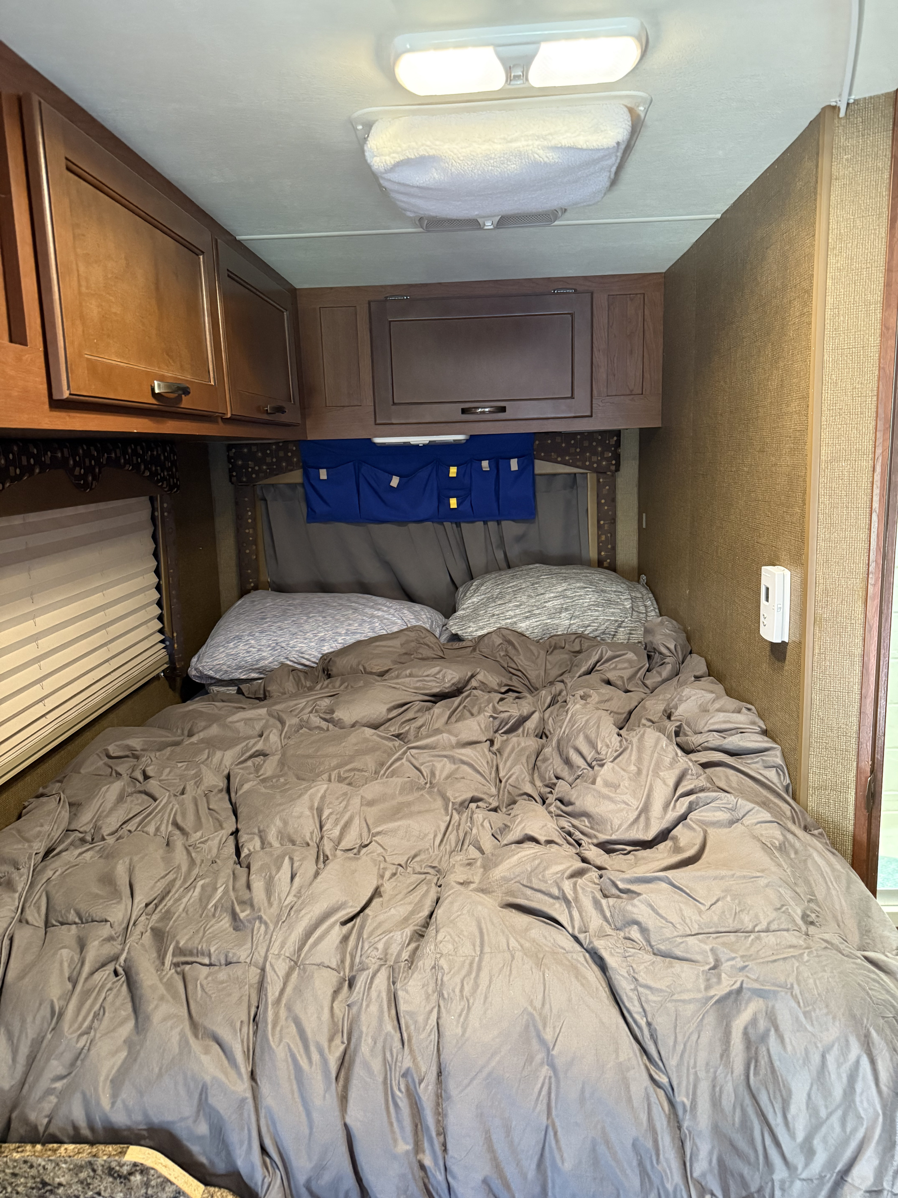 Inside an RV rental bedroom with a rumpled bed, overhead cabinets, ceiling light, and wall thermostat.. Thor Freedom Elite 2015
