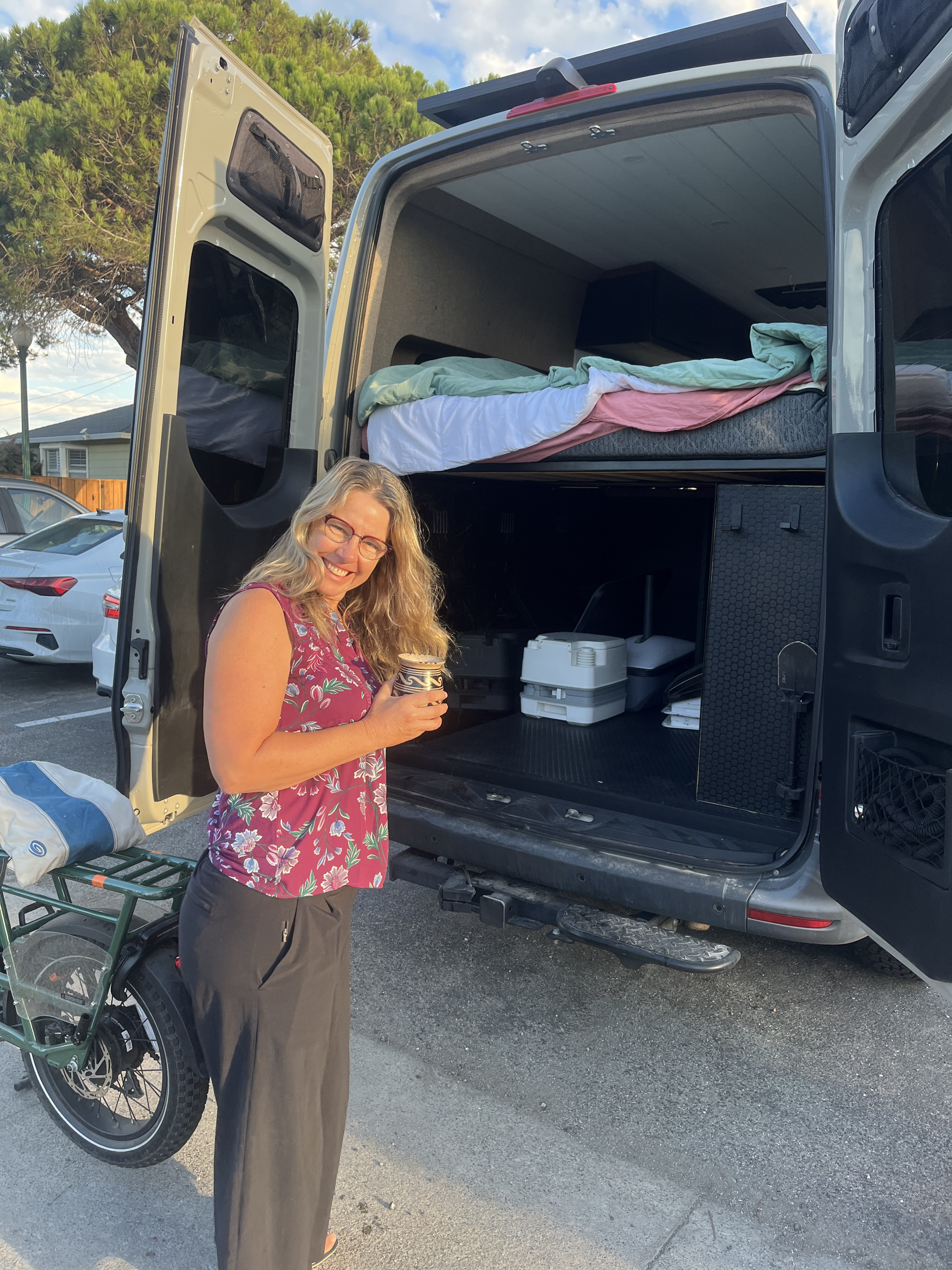 Smiling woman enjoys her RV rental camper van, revealing a cozy bed, portable toilet, and e-bike.. Mercedes-Benz Sprinter 2022