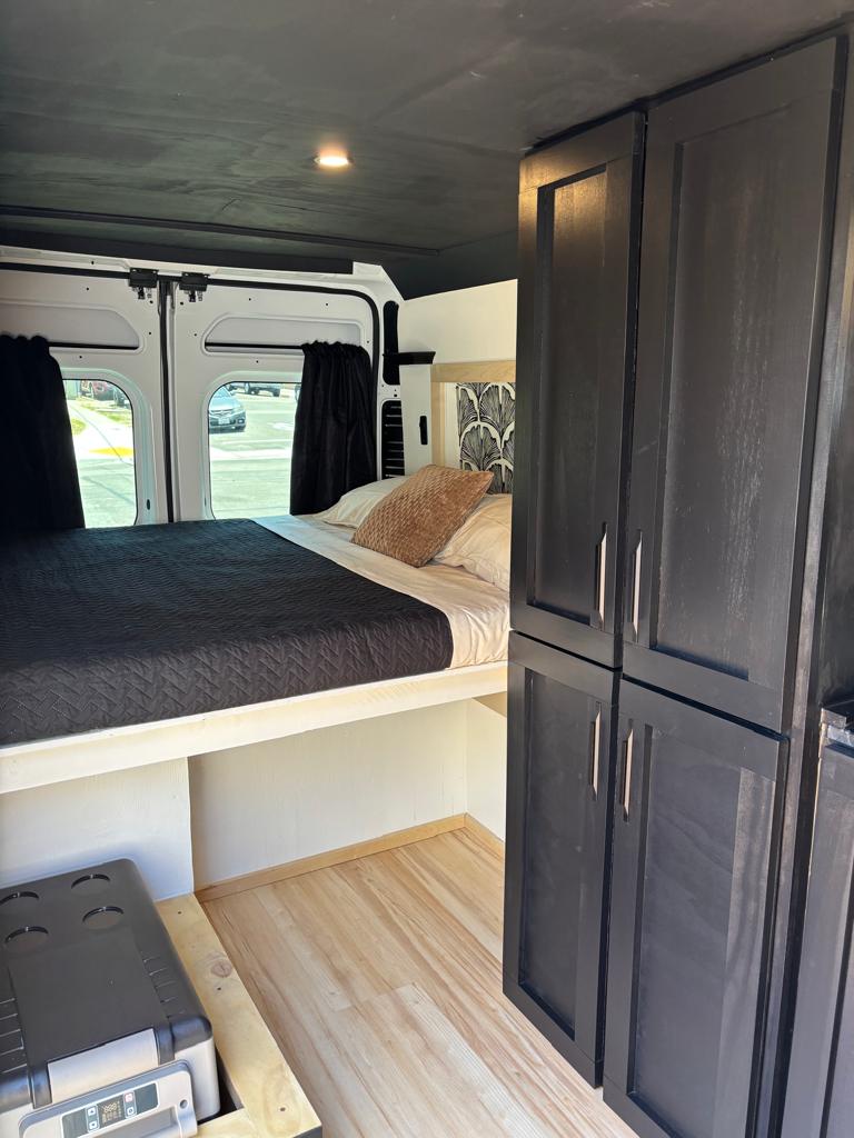 Step inside this RV rental's interior, featuring a cozy bed, dark storage cabinets, and a portable fridge.. Ram Promaster 2022