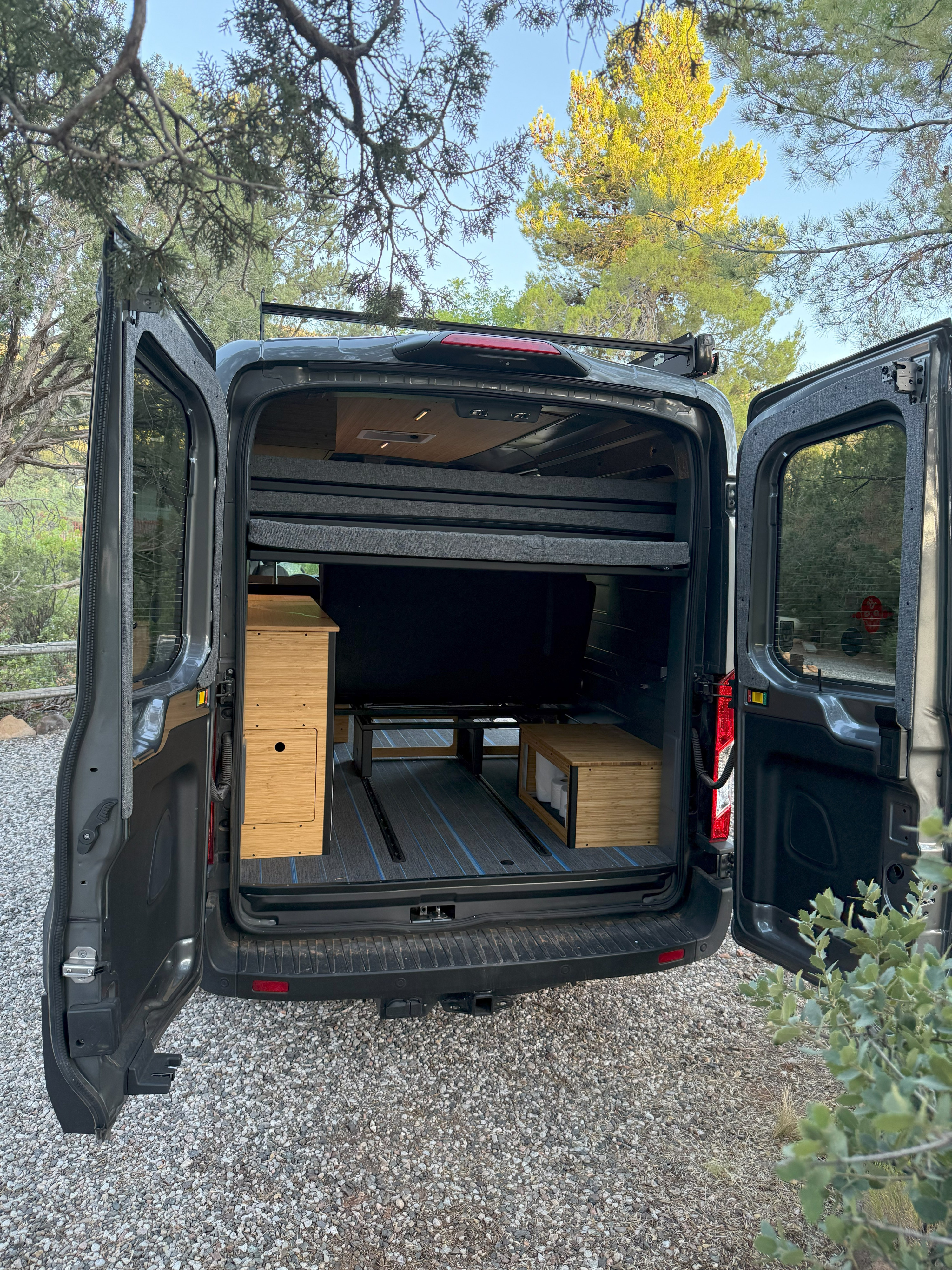 Explore the back of an RV rental camper van with open doors, showing its bed, wooden storage, and custom floor.. Ford Transit Custom 2024