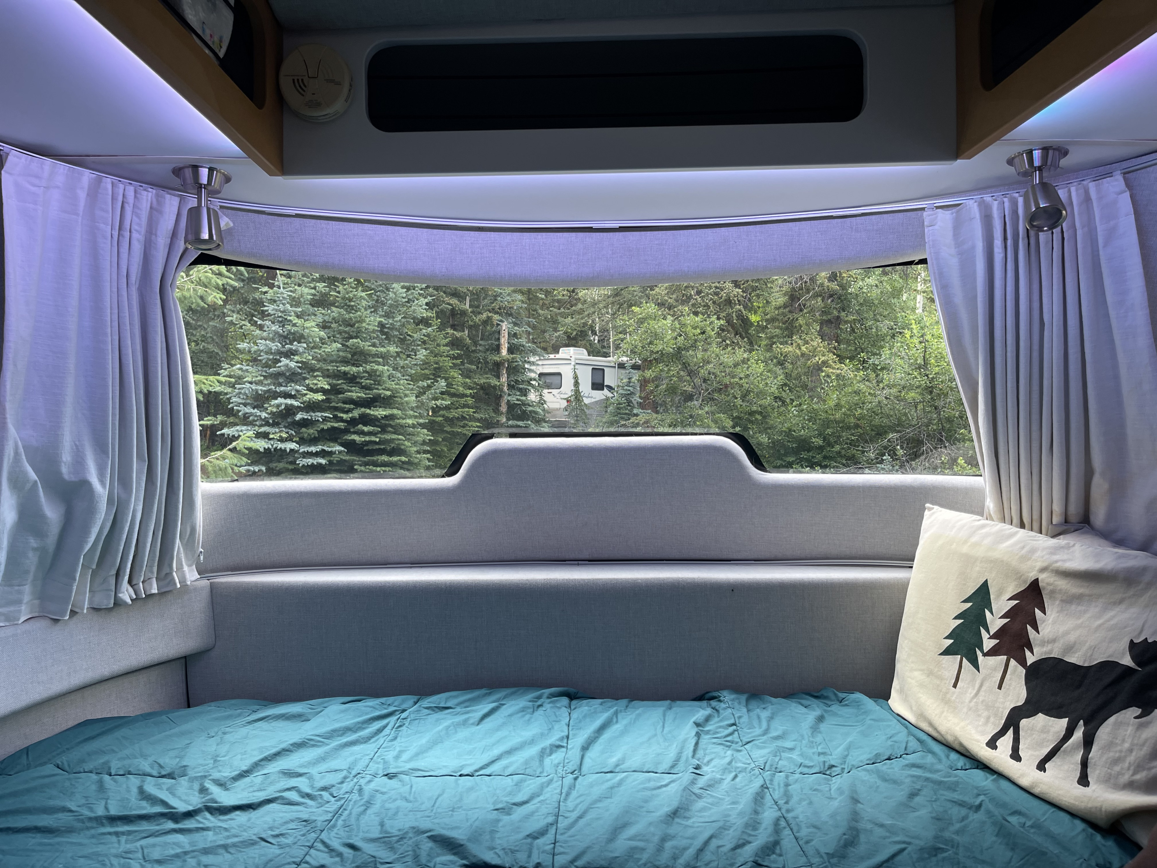 Serene RV rental bedroom looking out to a forest campsite with another RV visible. Teal bedding, grey interior.. Airstream Nest 2018