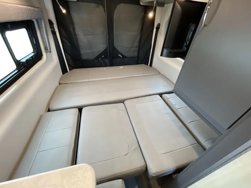 Inside an RV rental: light grey cushions form a bed in this cozy mobile living space. A window and TV are visible.. Thor Tranquility 2022