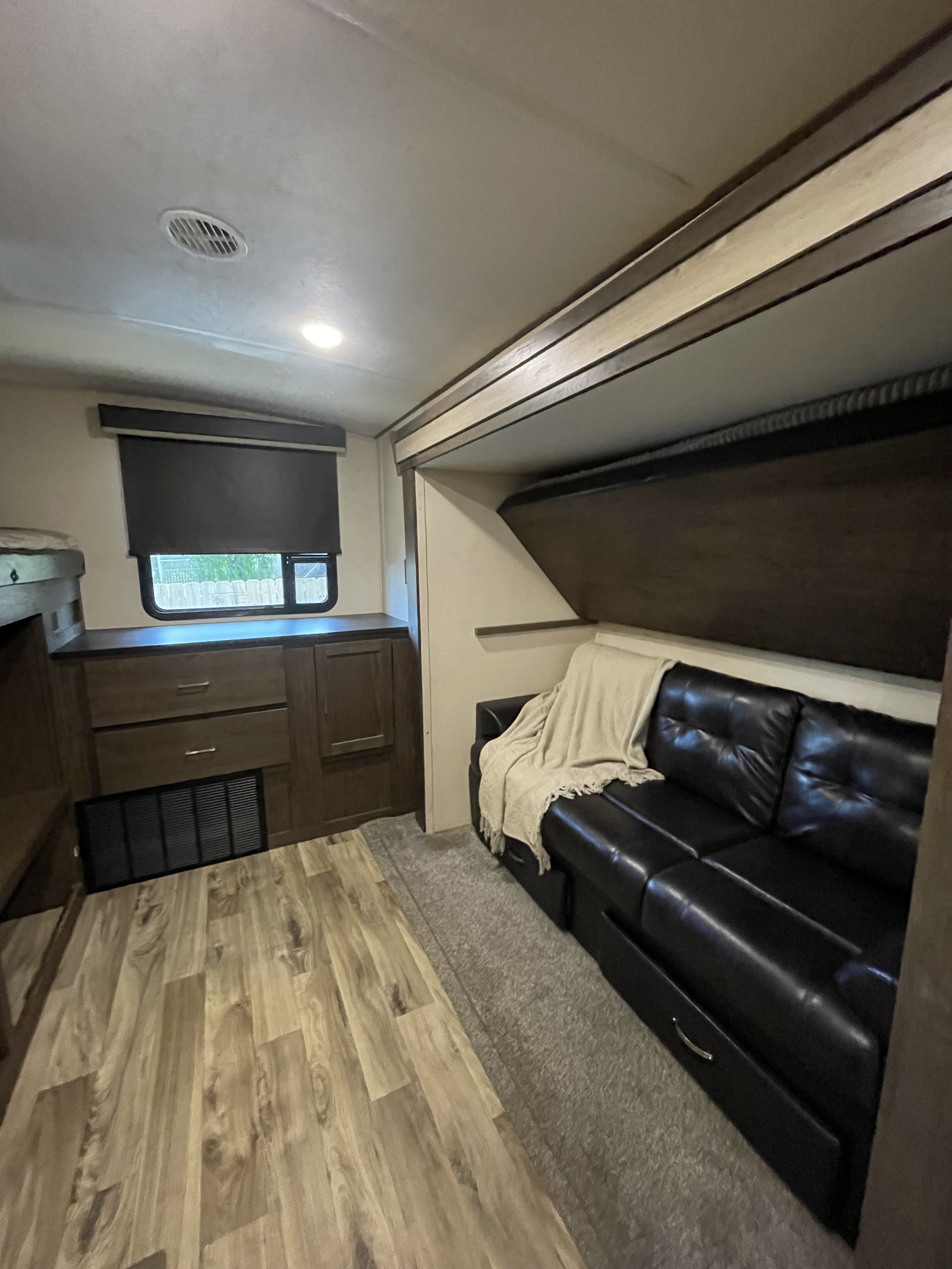 Inside an RV rental: living area with black leather couch, overhead bunk, wood-look floor, and window.. Forest River Wildwood Heritage Glen LTZ 2019
