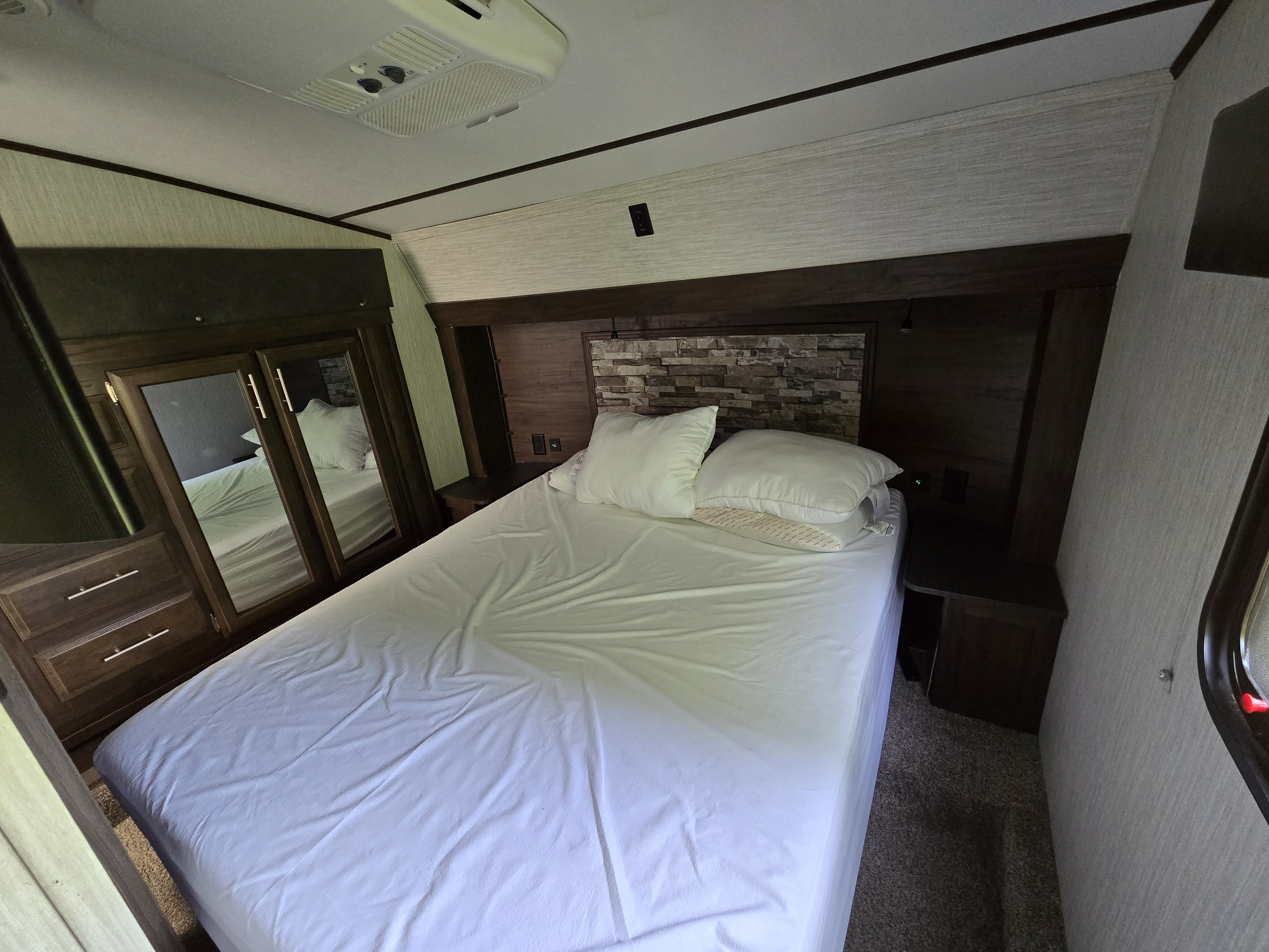 Inside a modern RV rental: a neat bedroom with a comfortable bed, stone headboard, mirrored wardrobe, and overhead AC.. Forest River Cherokee Arctic Wolf 2019