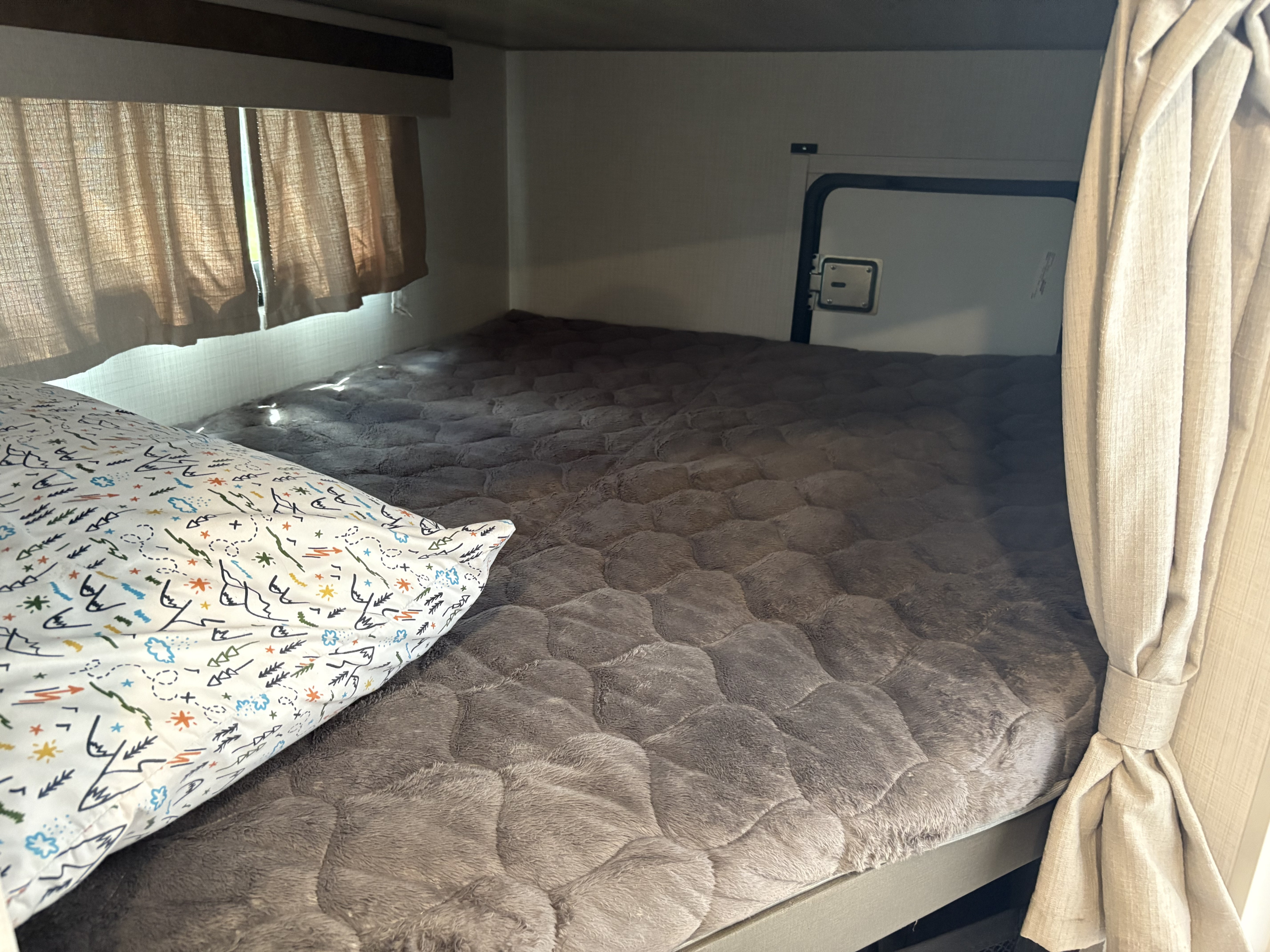 Inside an RV rental, a comfy bunk bed with a soft grey blanket, patterned pillow, and sunlight through curtains.. Forest River Rockwood Mini Lite 2024