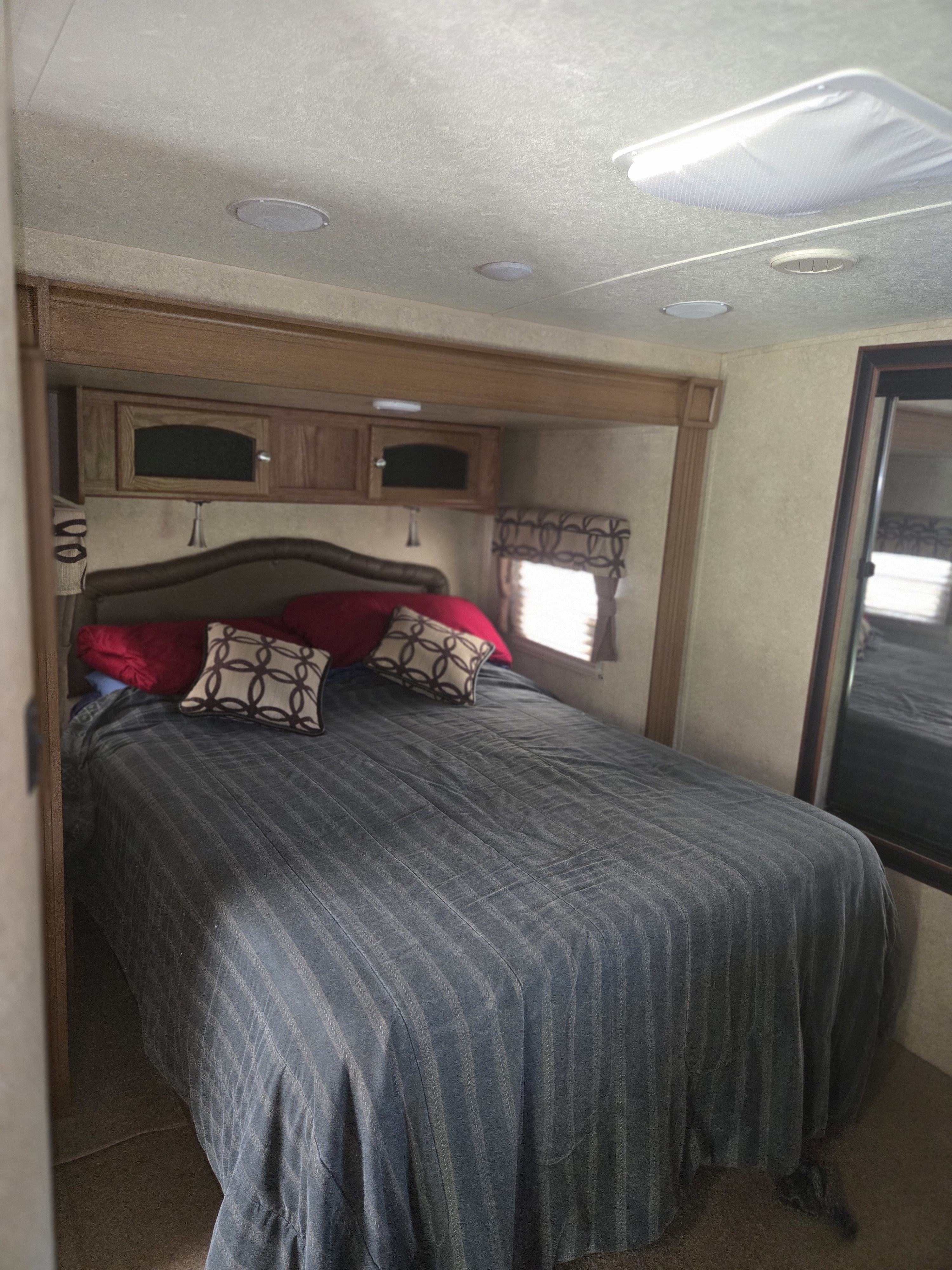 Cozy RV rental bedroom featuring a full bed with red and patterned pillows, overhead cabinets, and a side window.. Forest River Flagstaff Classic Super Lite 2016