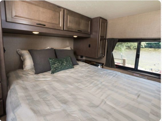RV rental bedroom with comfortable bed, overhead cabinets, and a scenic window view of water.. Thor Majestic 2019