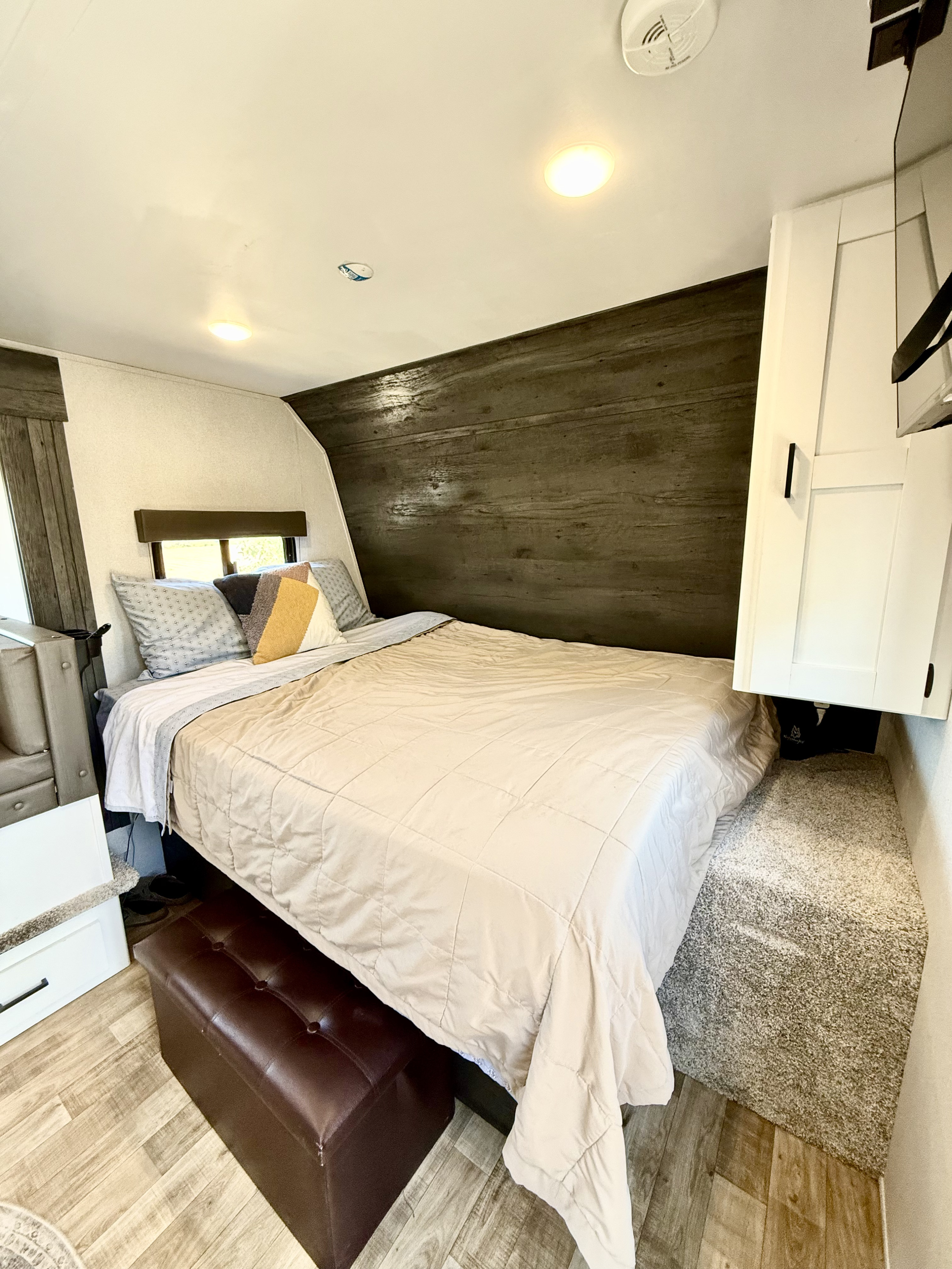 View of an RV rental bedroom with a comfortable bed, dark wood accent wall, overhead cabinets, and warm lighting.. Keystone RV Hideout 2022