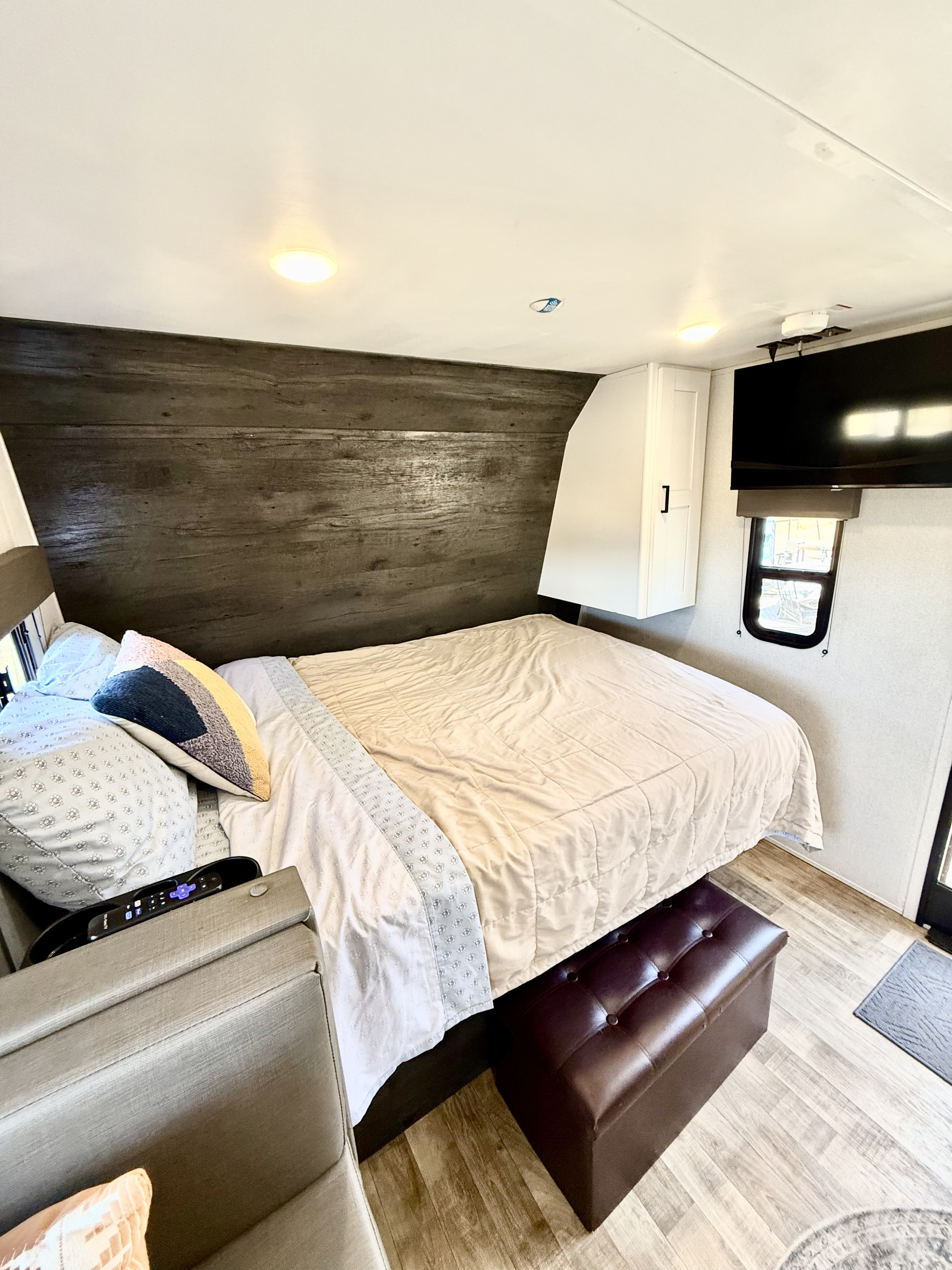 Cozy RV rental bedroom with a large bed, dark wood accent wall, wall-mounted TV, cabinet, ottoman, and wood-look floor.. Keystone RV Hideout 2022