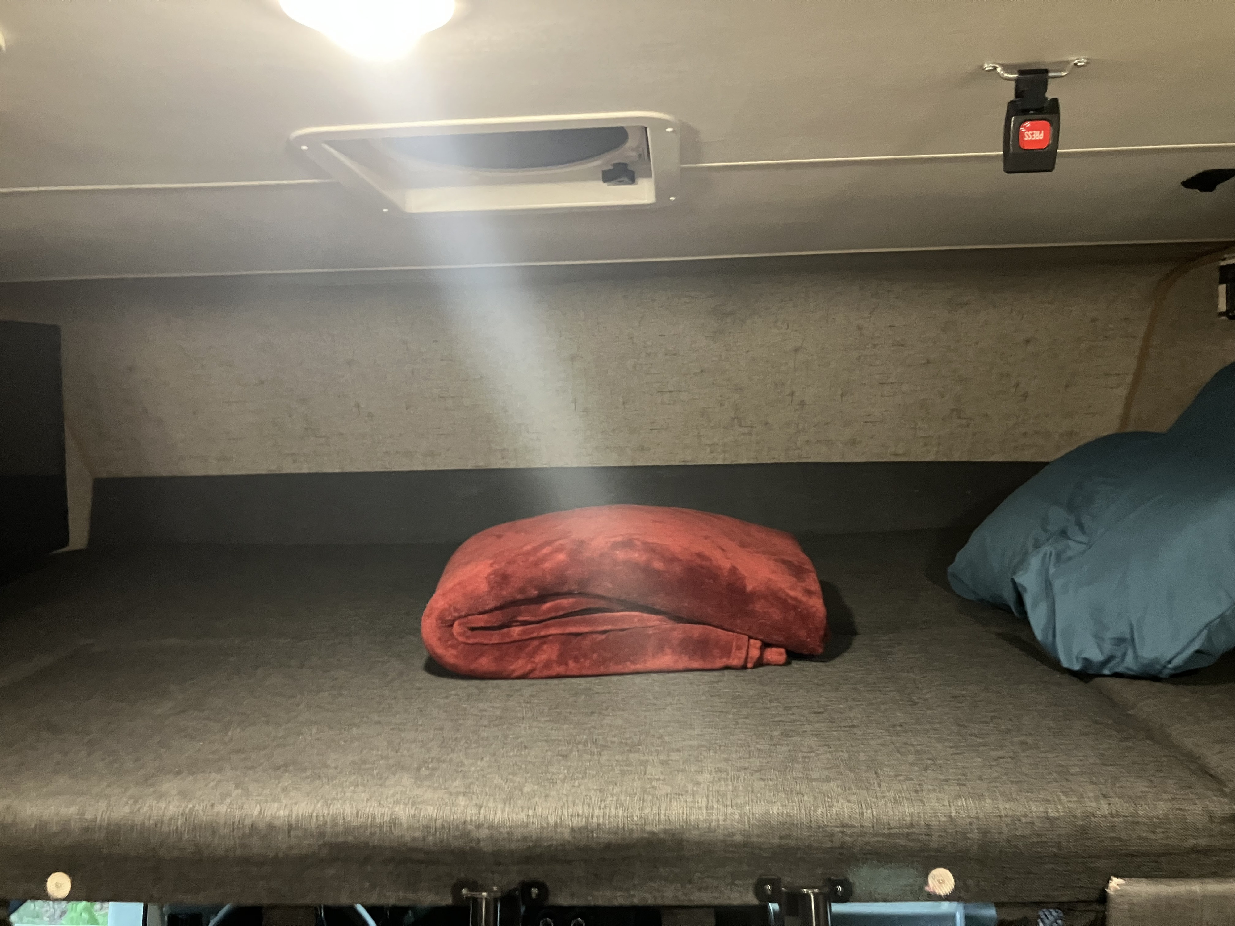 This RV rental's cab-over bunk features a comfy bed with a red blanket, blue pillow, ceiling vent, and safety buckle.. Forest River Coachmen Freelander 2017