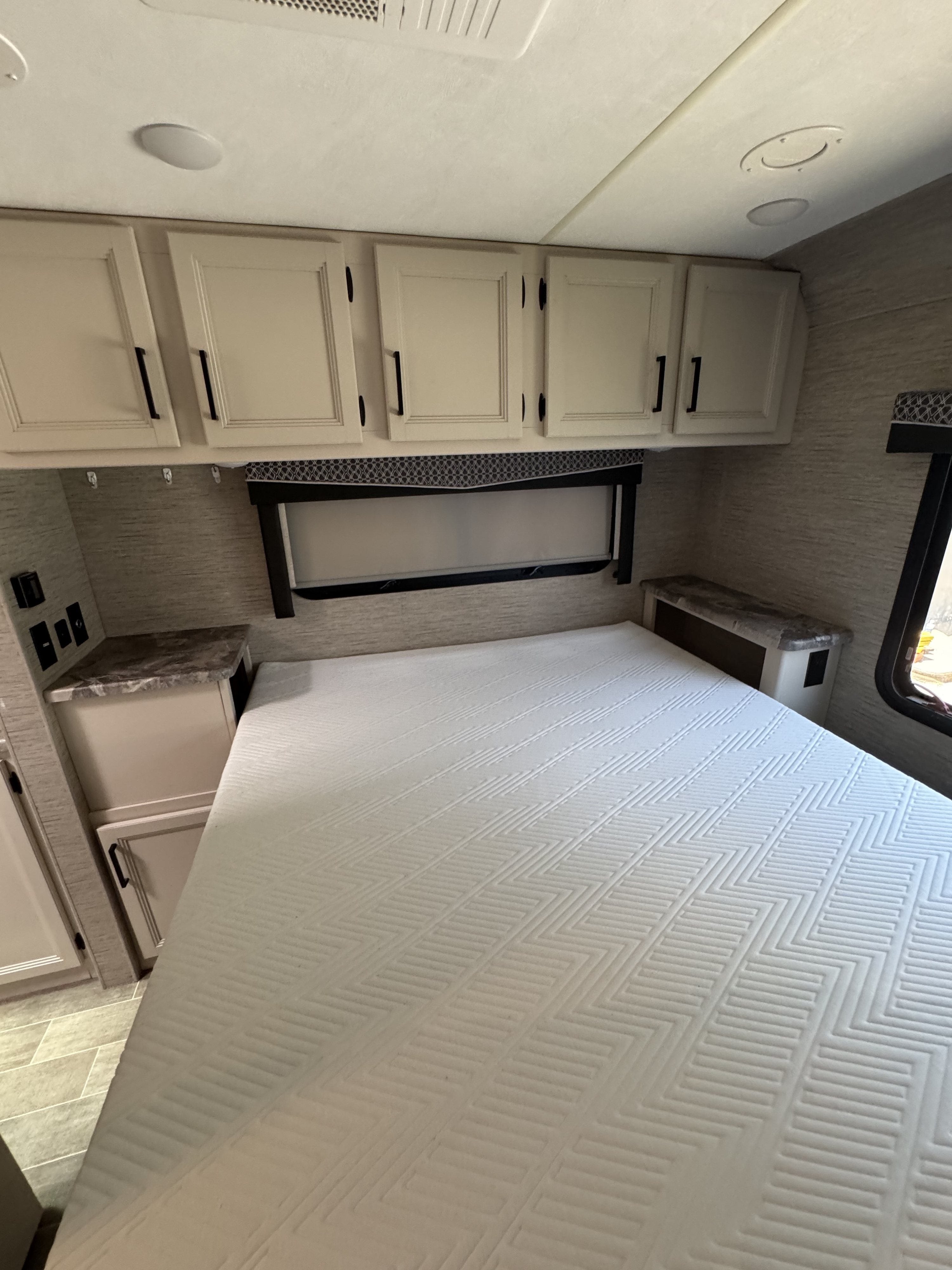 An RV rental's inviting bedroom. Features a white patterned mattress, light cabinets, bedside outlets, and a window.. Four Winds Chateau 2022