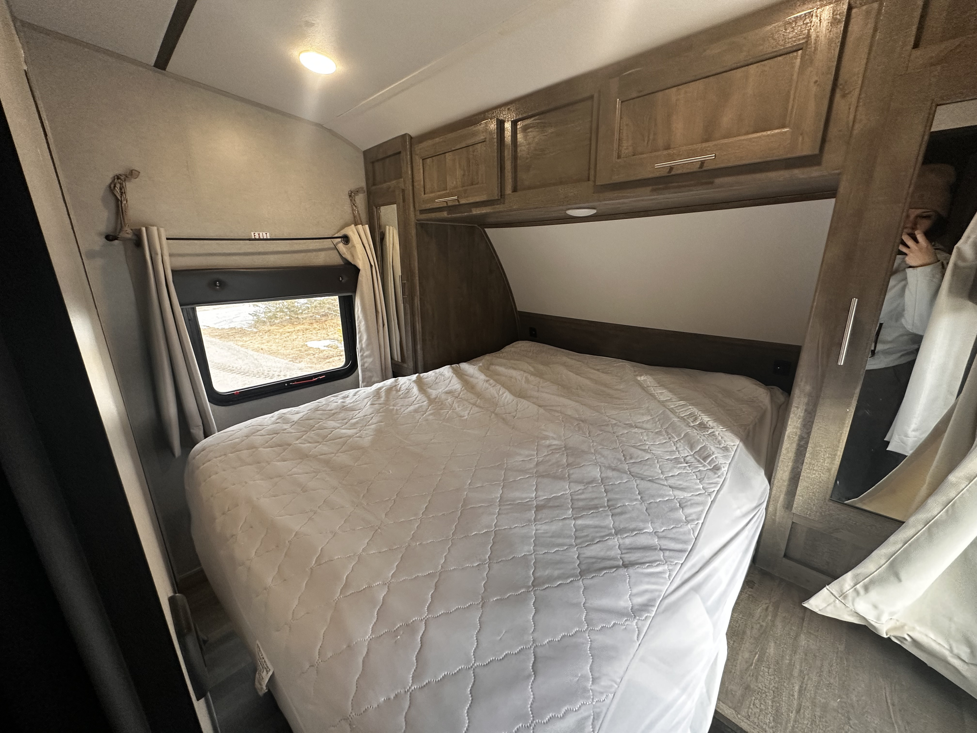 Cozy RV rental bedroom featuring a quilted bed, wooden overhead cabinets, and a window with an "EXIT" sign.. Forest River XLR Boost 29QBS 2020