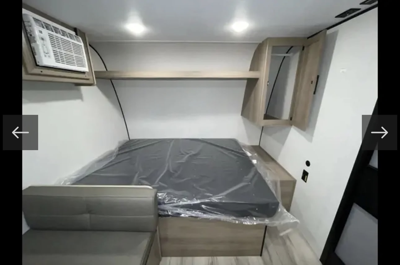 RV rental interior: A bedroom with a plastic-covered mattress, wall-mounted AC, and overhead storage.. KEYSTONE COLEMAN 17B 2025