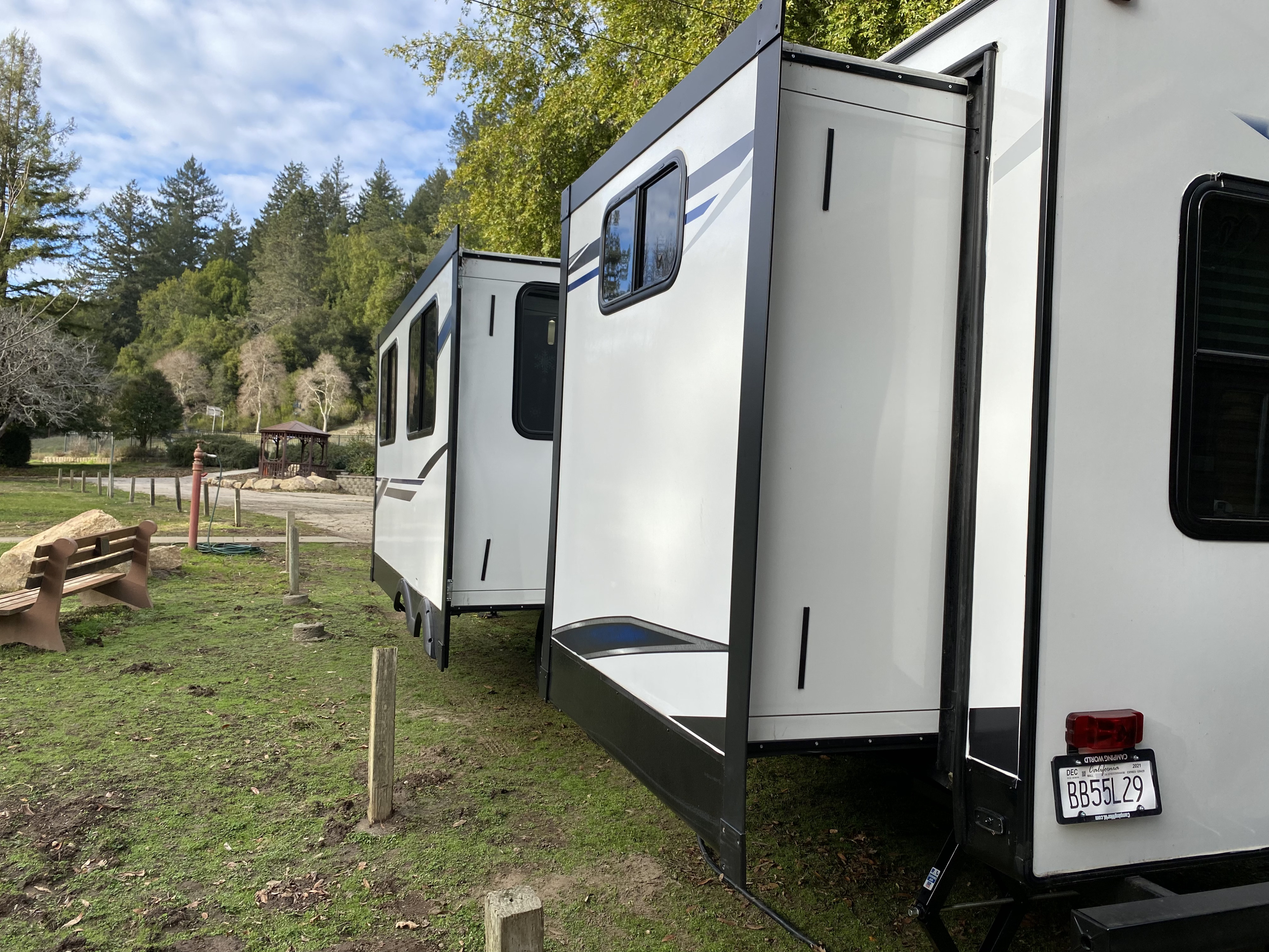 White RV rental with extended slide-outs parked at a green campsite, surrounded by trees and a gazebo.. Heartland RVs Mallard 2021