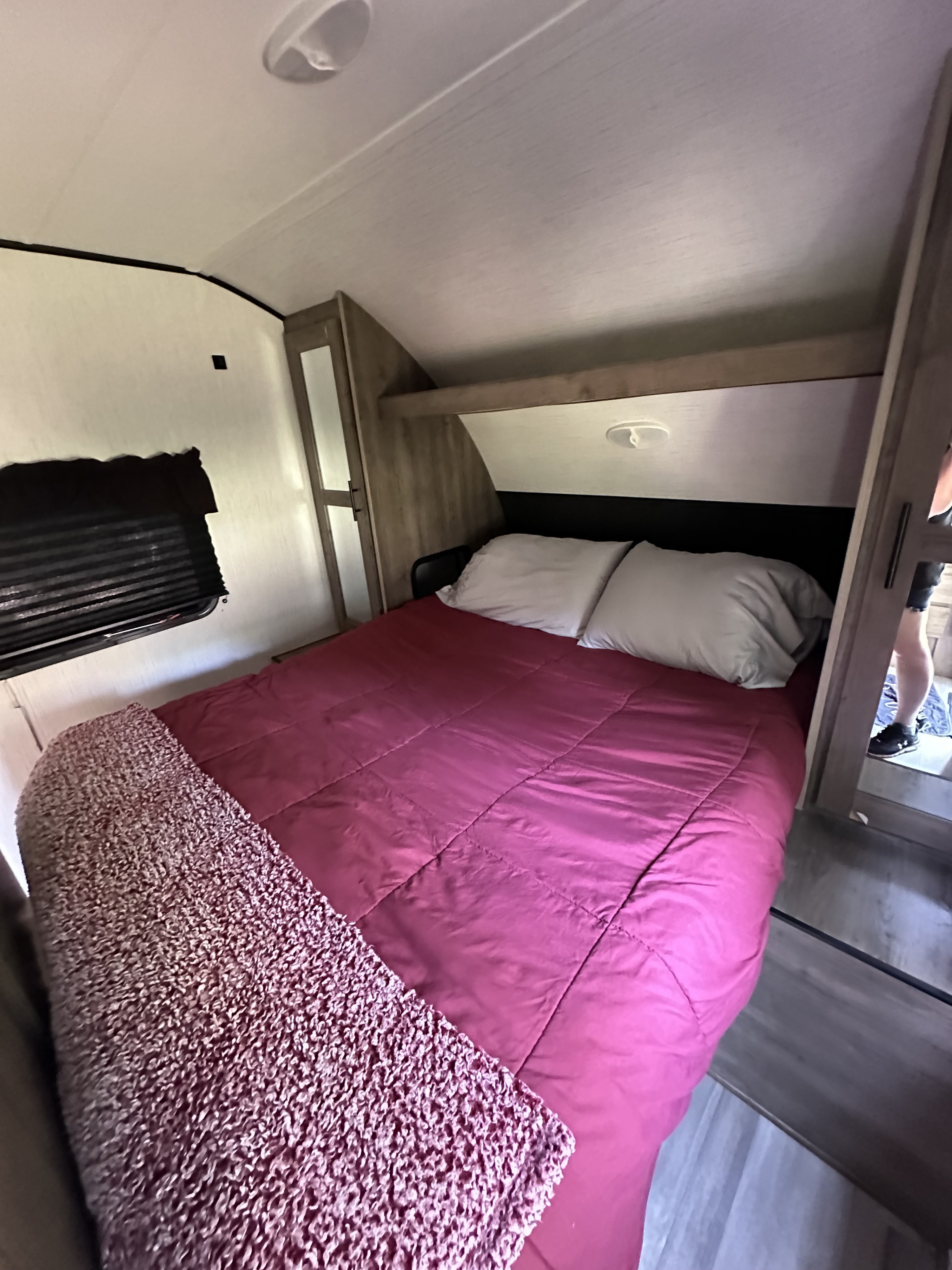 Cozy RV rental bedroom featuring a bed with a red comforter, white pillows, and a window with blinds.. Heartland RVs Pioneer 2022