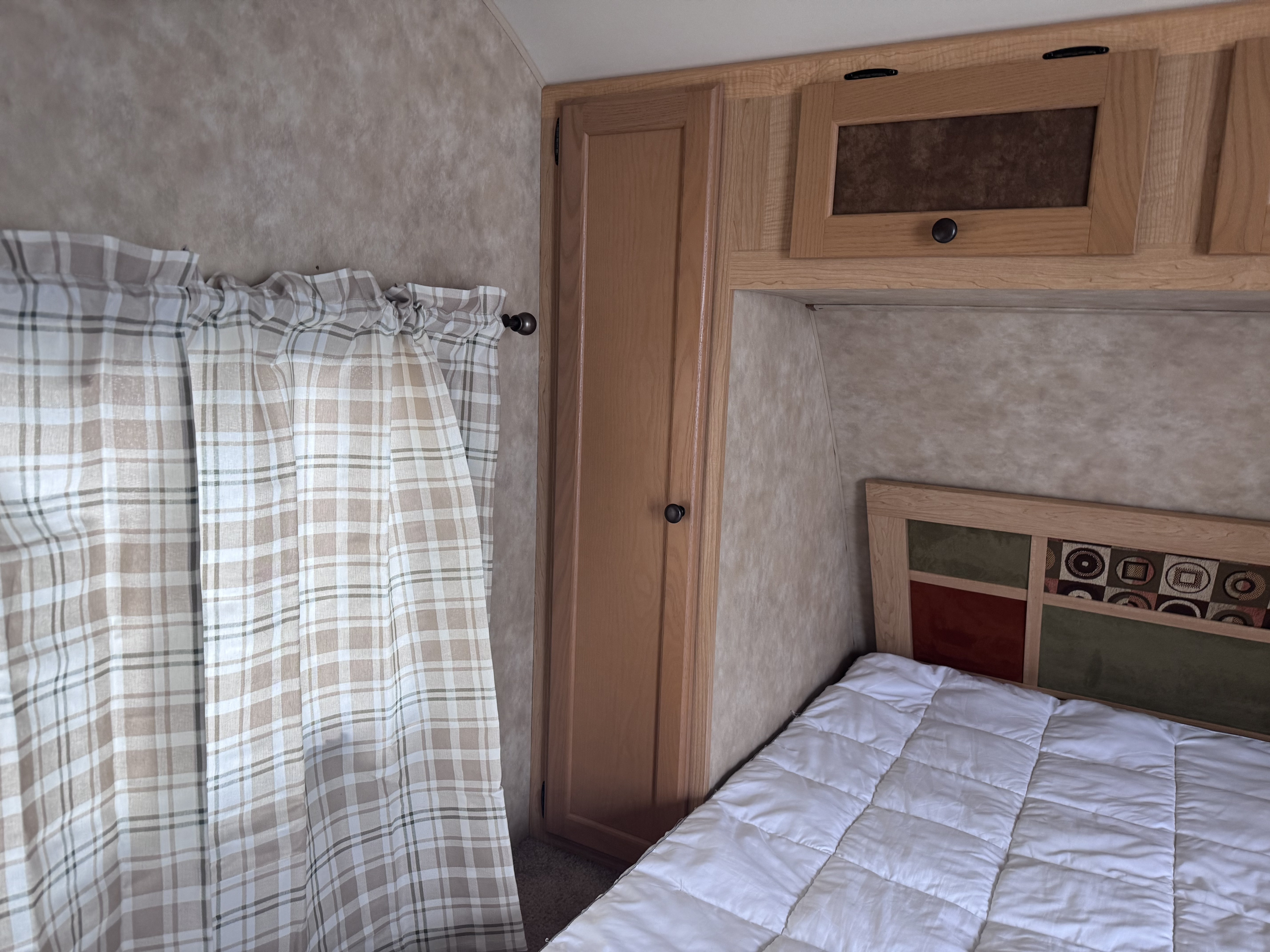 RV rental bedroom: plaid curtains, quilted bed with decorative headboard, and wooden storage cabinets.. kz Coyote 2010