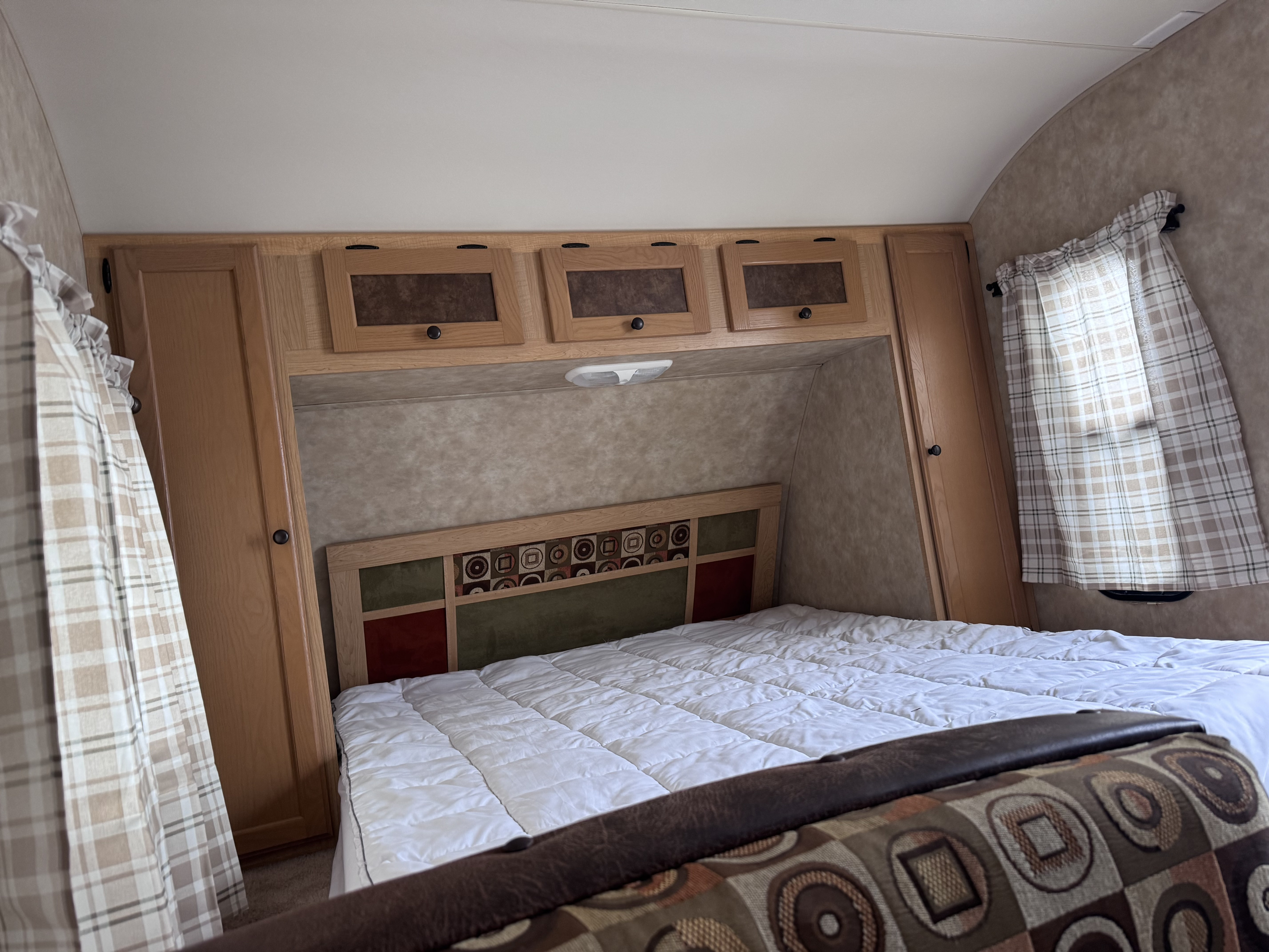 RV rental bedroom: A cozy space with a white bed, patterned headboard, overhead wooden cabinets, and plaid curtains.. kz Coyote 2010