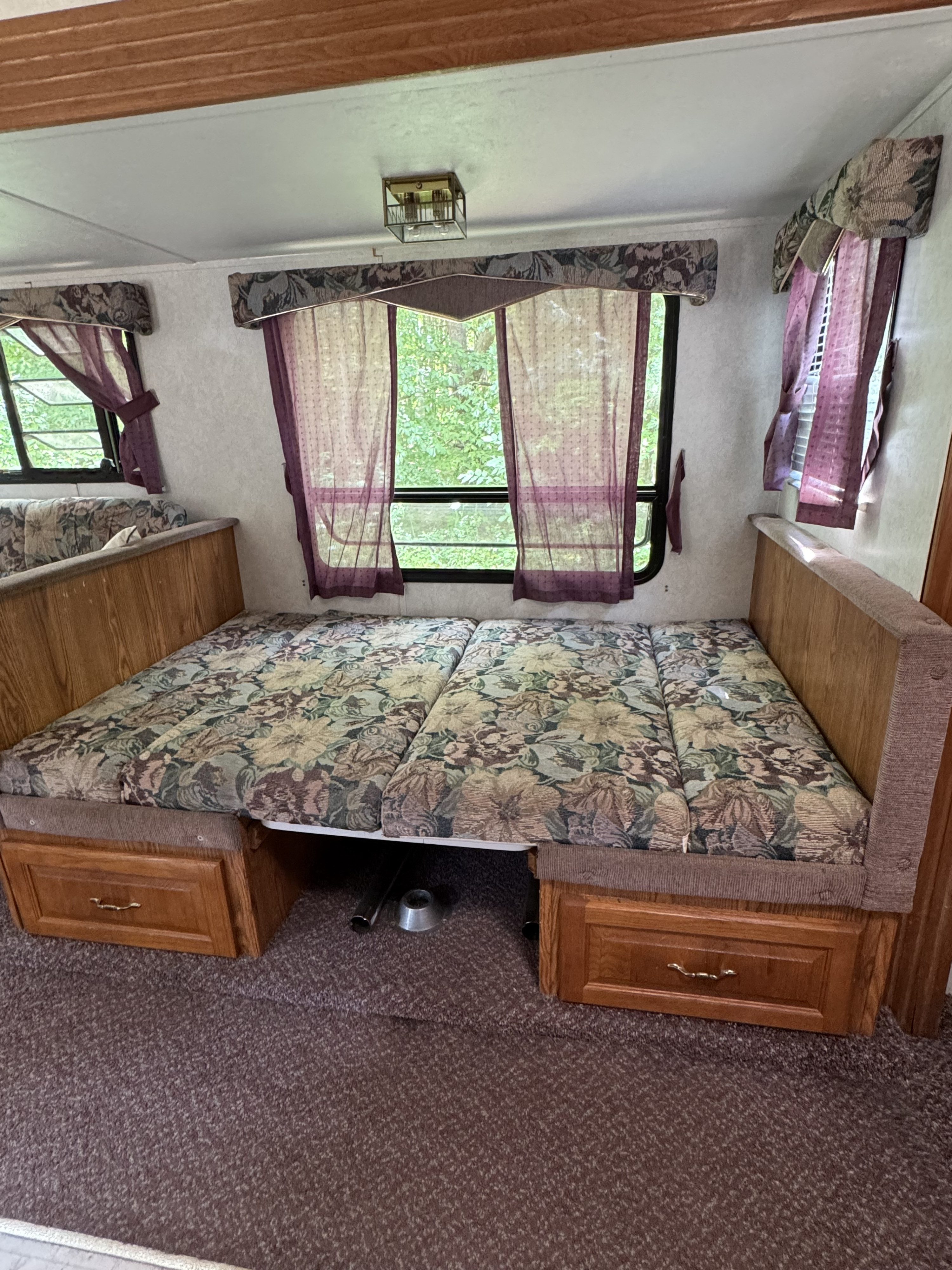 RV rental interior featuring a convertible floral dinette bed, wood storage, and window with purple curtains.. Forest River Wildwood 2000