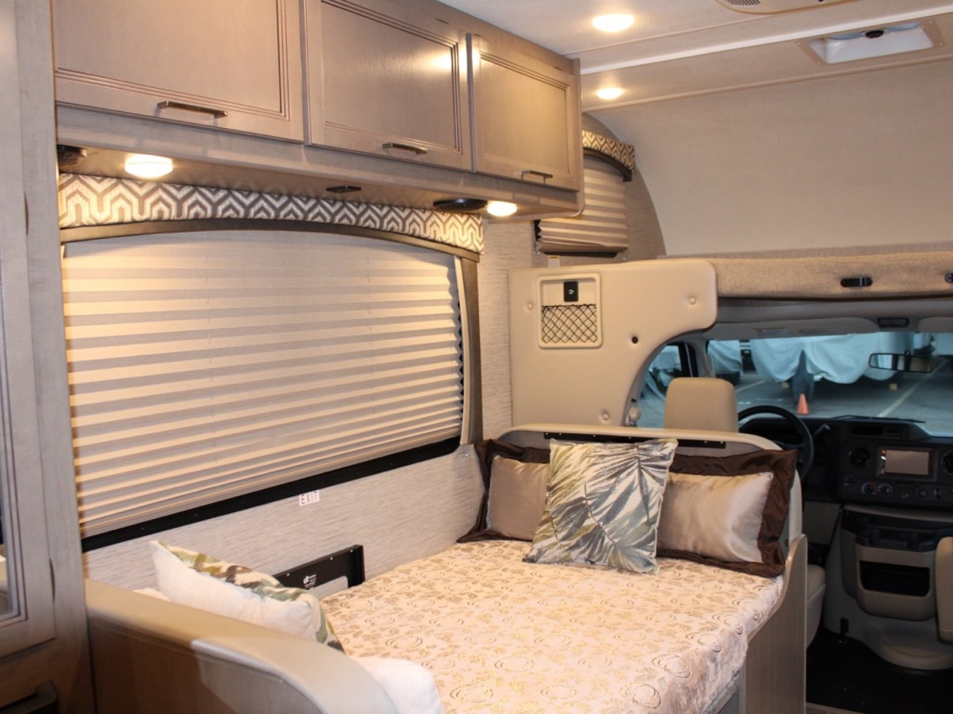 Cozy rv rental interior: a comfortable bed with patterned bedding, overhead cabinets, and front driving area.. Thor Chateau 2022