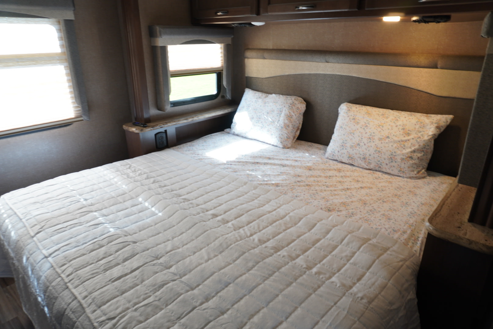 Cozy RV rental bedroom featuring a comfortable bed with a white quilted comforter and floral pillows, plus two windows.. Thor A.C.E. 2018