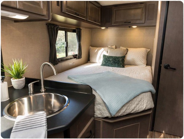 Cozy RV rental interior: a comfortable bed with a light blue throw, a compact kitchenette featuring a sink, and practical overhead cabinets.. Thor Majestic 2019