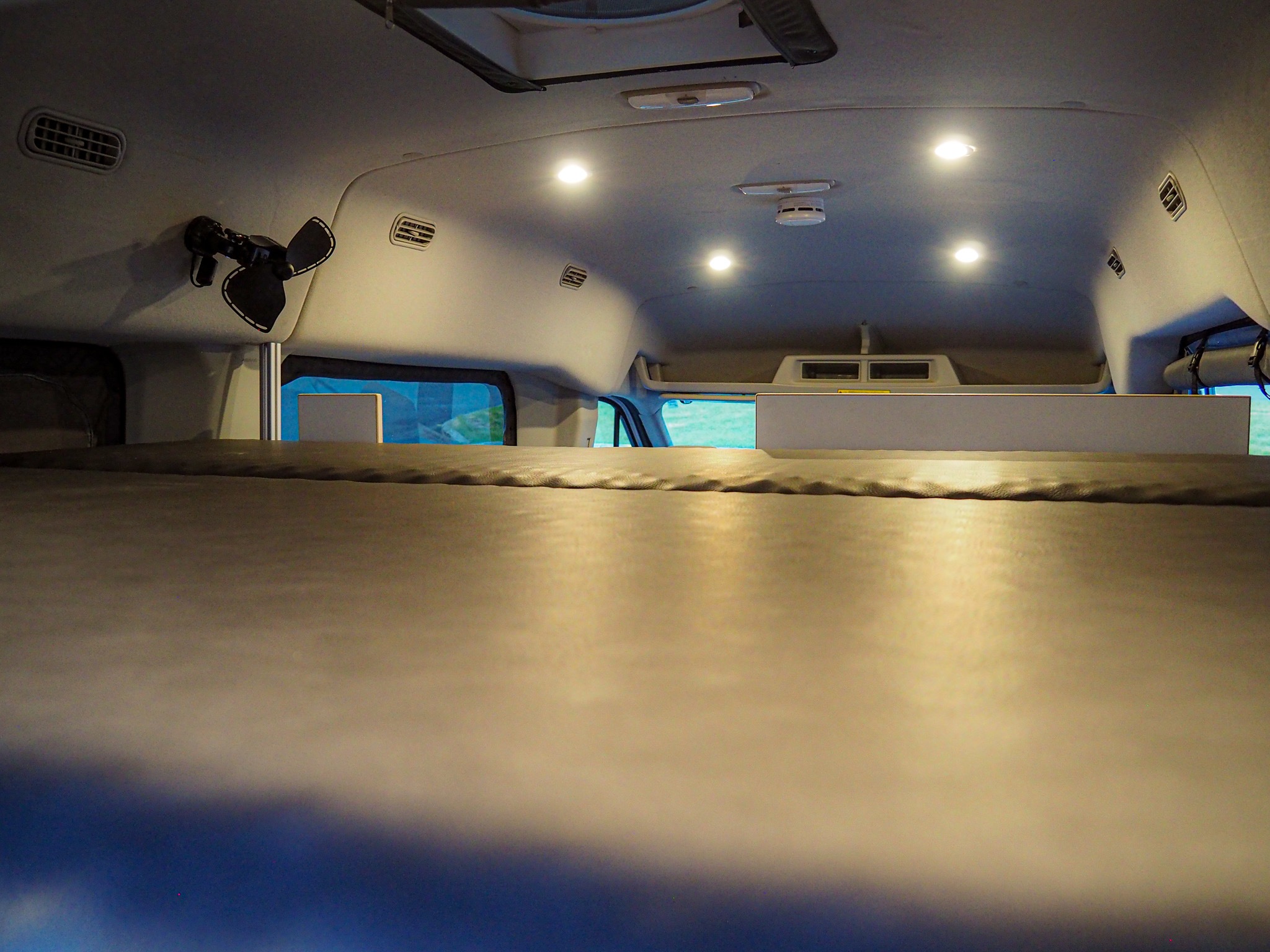 Inside an RV rental van, looking forward at the bed and well-lit ceiling with recessed lights, air vents, and a fan.. Ford Transit 2019