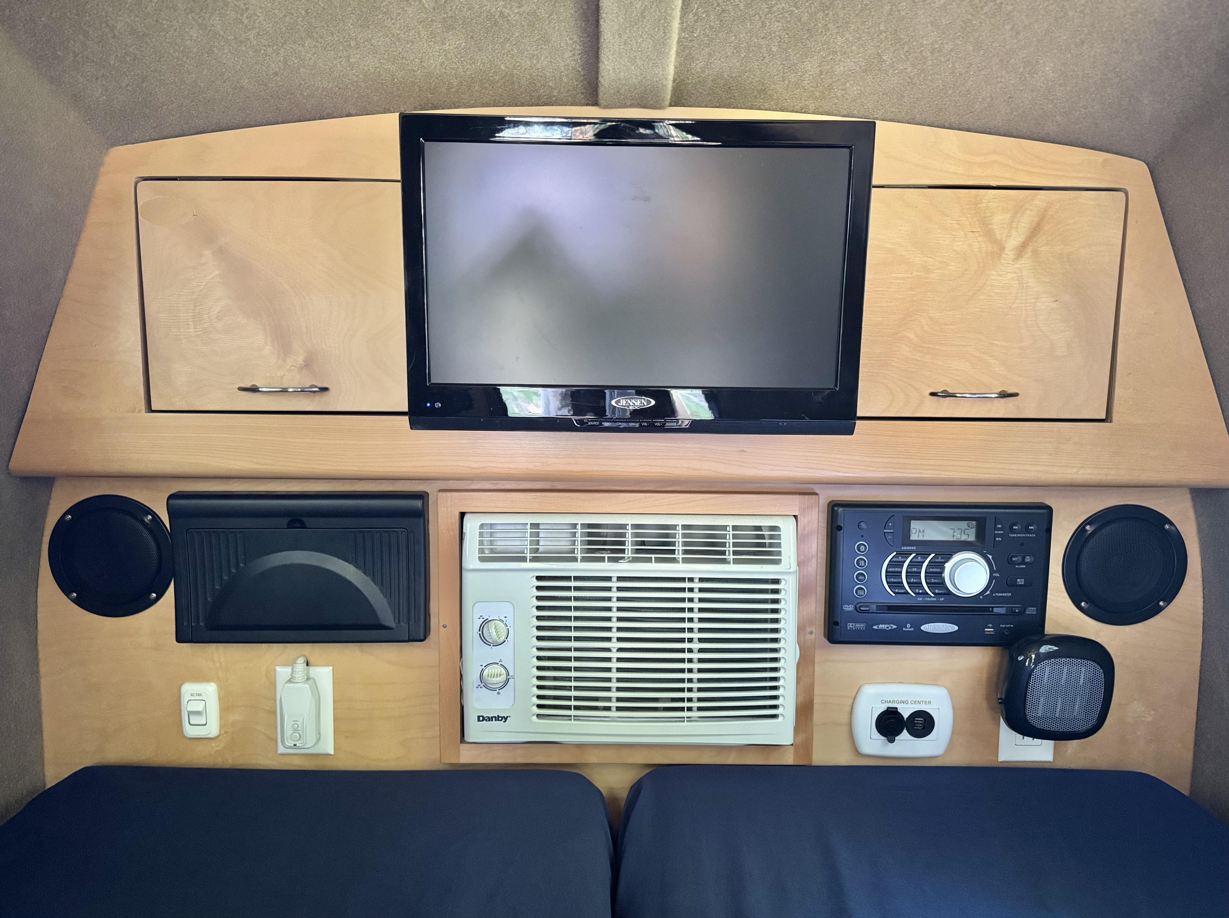 Interior of an RV rental: TV, AC unit, stereo, speakers, charging center, and other controls built into light wood cabinetry.. Little Guy MyPod 2017