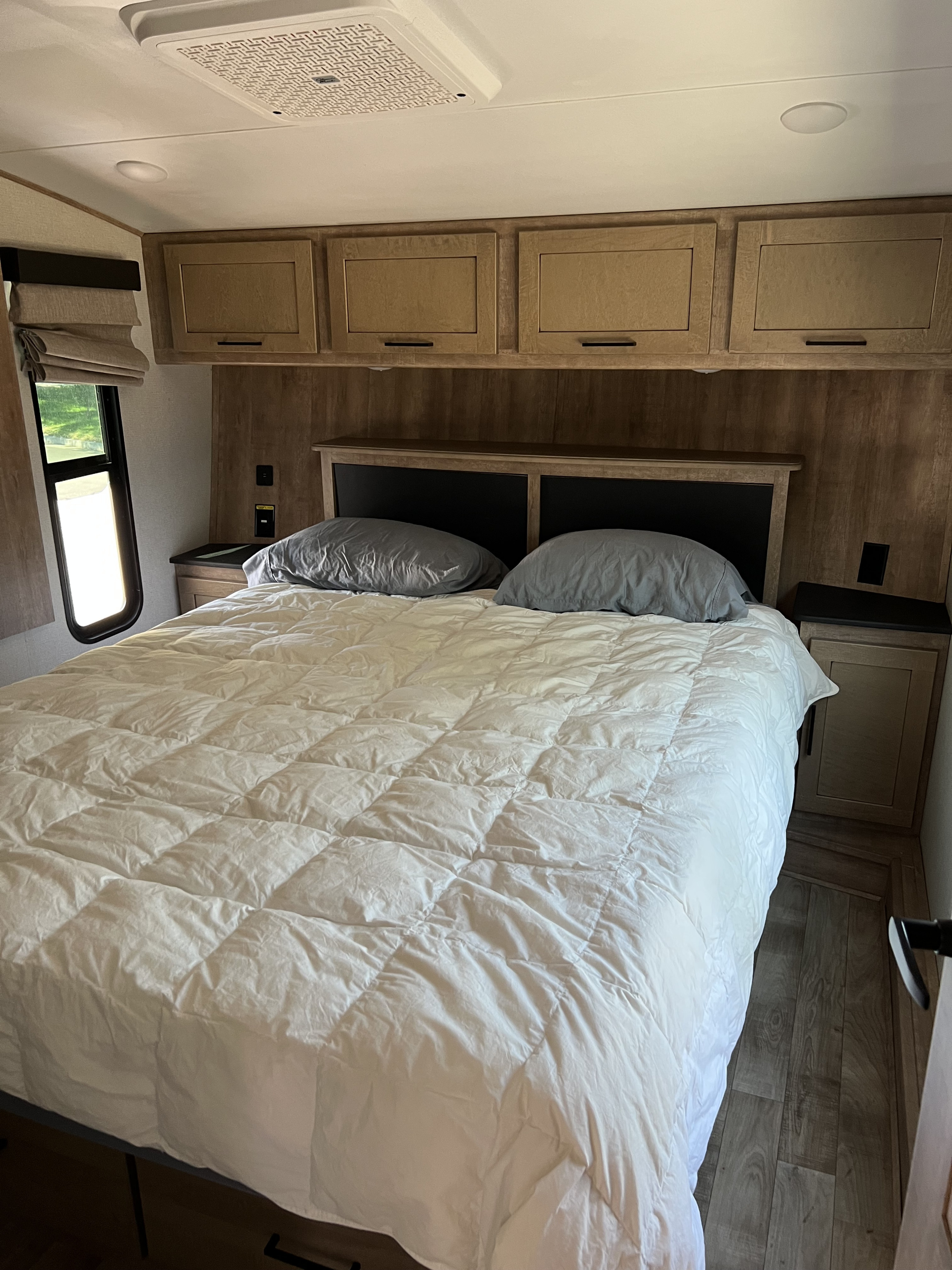 A cozy RV rental bedroom showing a large bed, white comforter, wood cabinets, nightstands, and ceiling AC unit.. Keystone RV Arcadia 2022