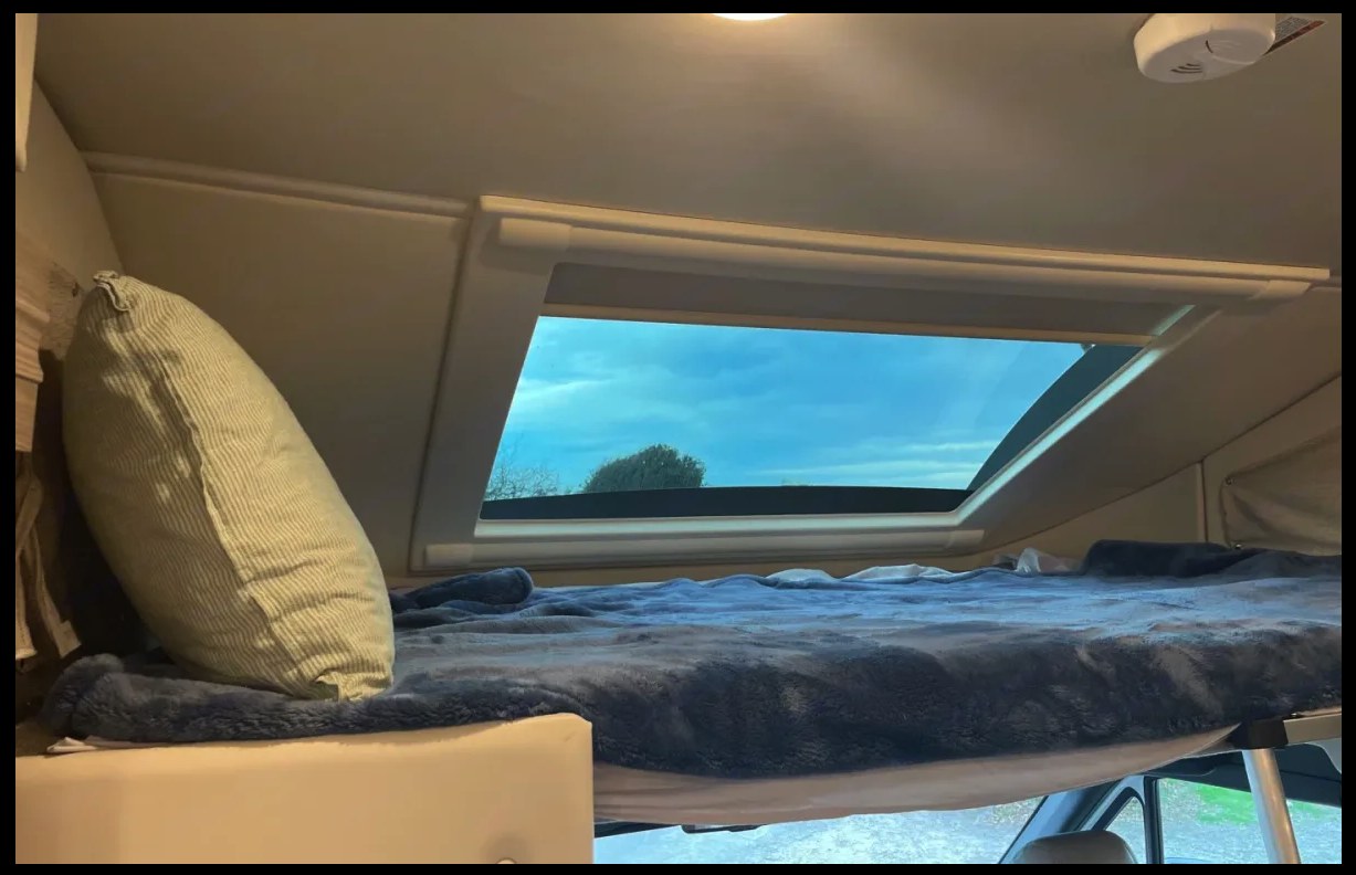 An RV rental's cozy overcab bunk bed with a blue blanket, pillow, and a large skylight showing a blue sky. Ideal for travel.. Thor Motor Coach Delano 2022