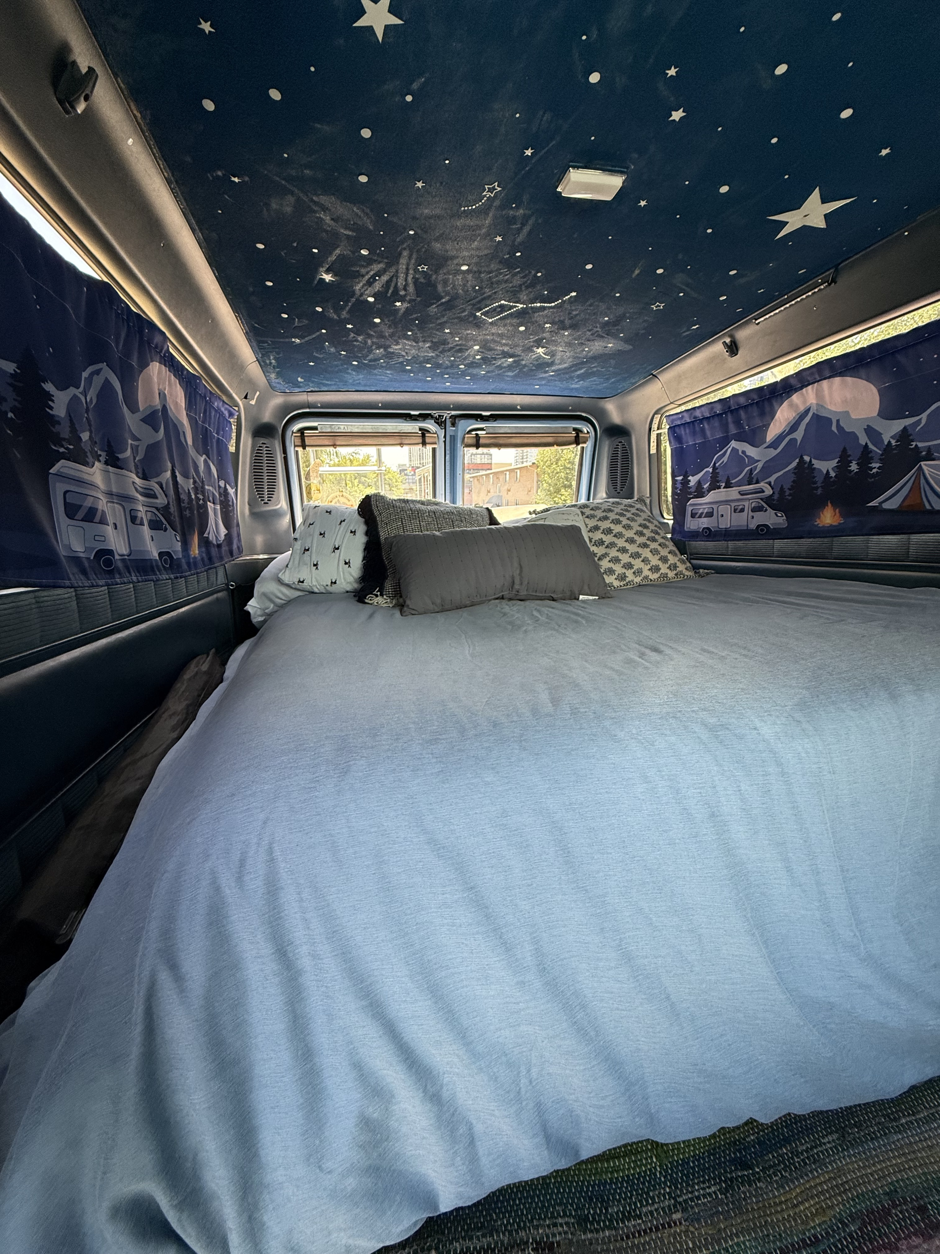 Inside a spacious rv rental: a cozy bed with pillows, starry ceiling, and nature-themed curtains.. Gmc Rally Wagon 1985