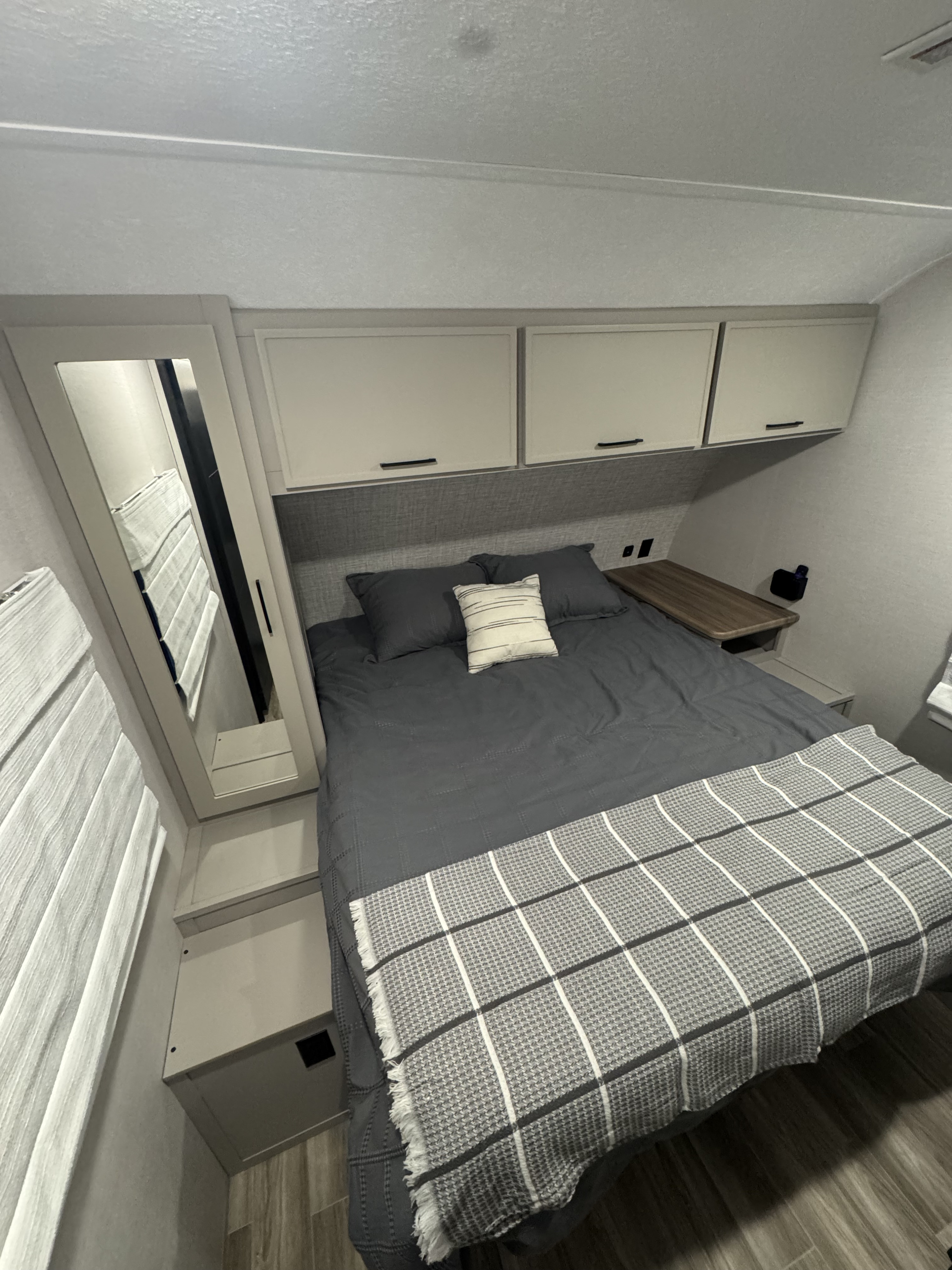 Cozy RV rental bedroom features a grey bed with plaid throw, overhead cabinets, mirror, and compact design.. Forest River Vibe 2024