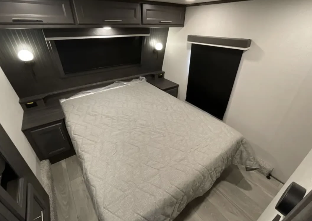 This RV rental bedroom offers a comfortable bed, dark overhead cabinets, nightstands, and a window with a blind.. Keystone Cougar 2025