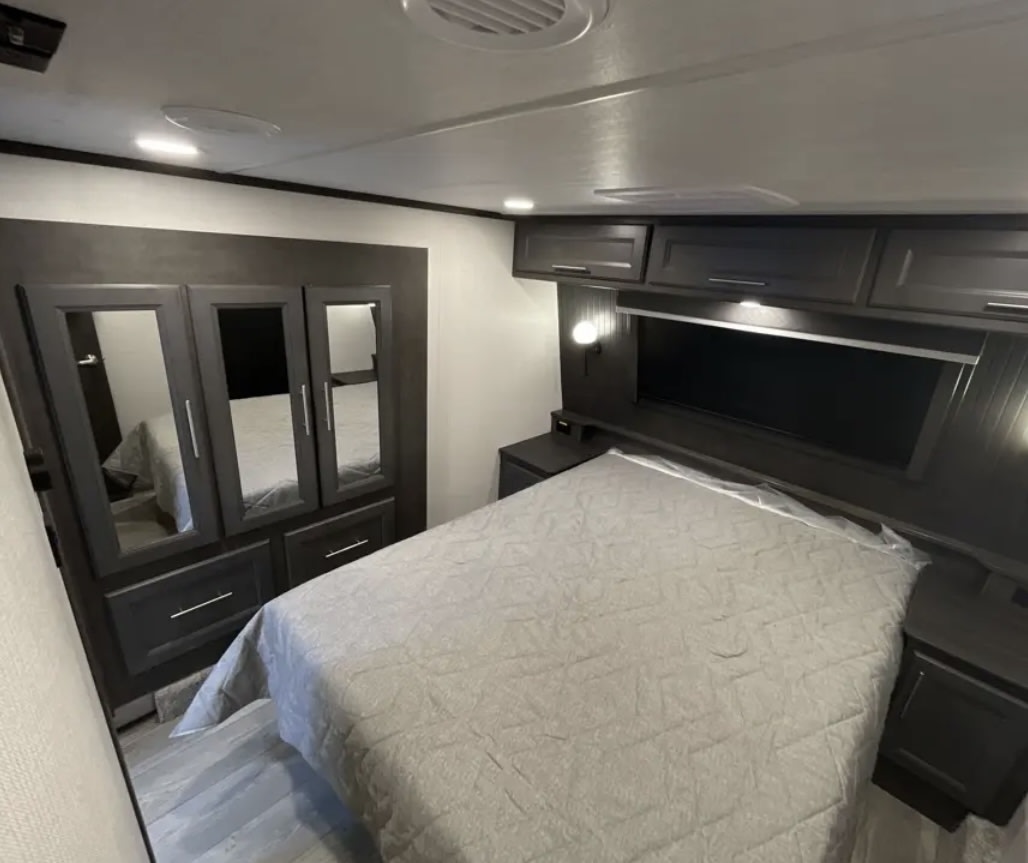 Step into this modern RV rental bedroom: a comfortable quilted bed, dark wood cabinets, and mirrored wardrobe storage.. Keystone Cougar 2025