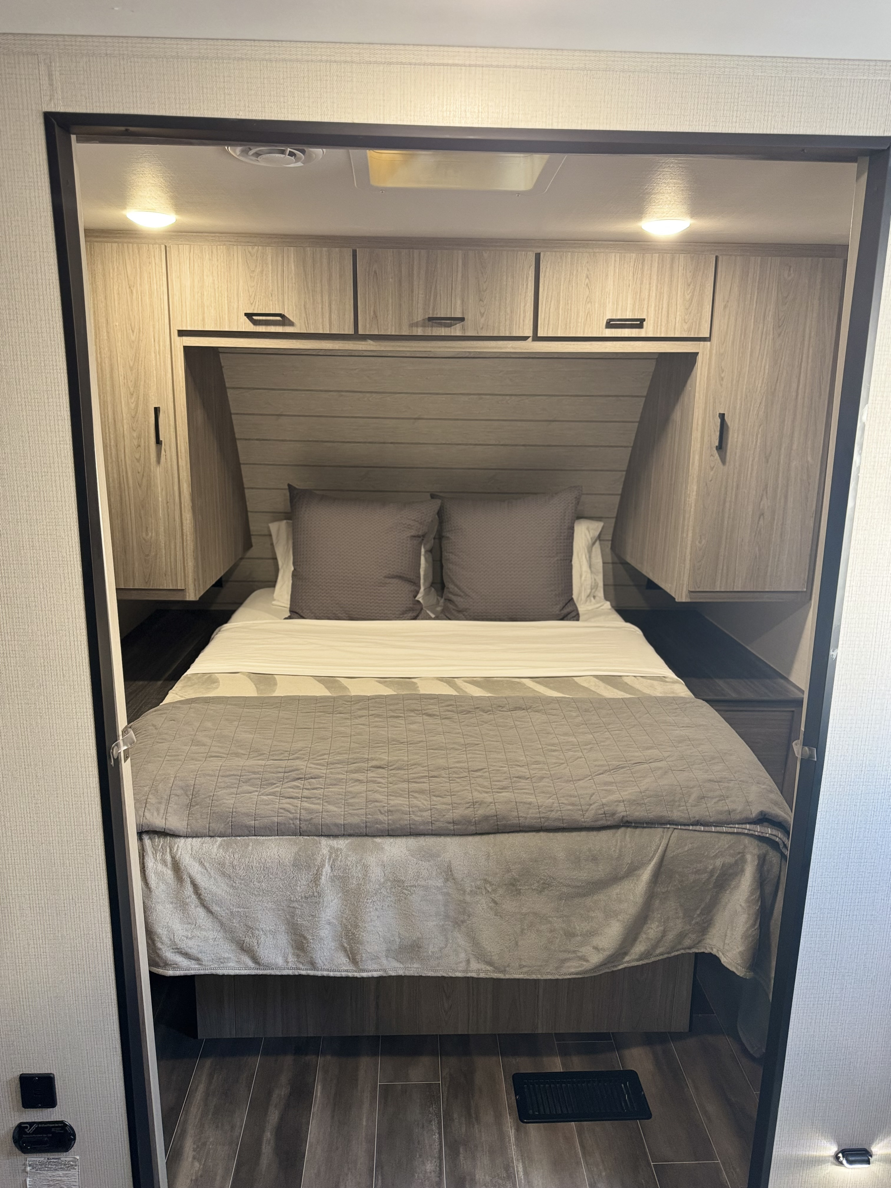 Inside an RV rental, this cozy bedroom features a bed with grey bedding, overhead cabinets, and wood-look flooring.. Keystone RV Passport TT 2022